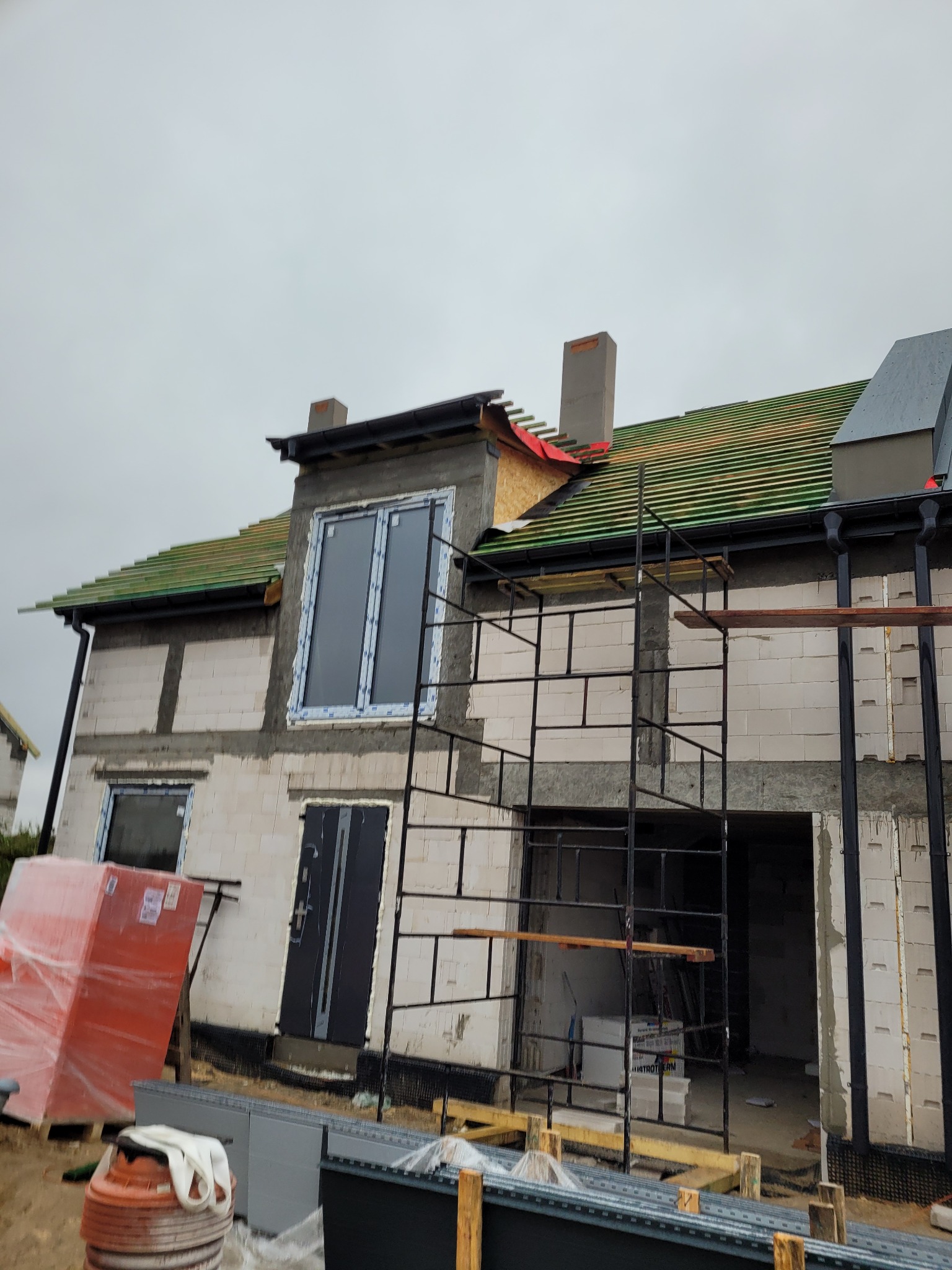 Budowa domu jednorodzinnego w stanie surowym otwartym, widoczne rusztowania, częściowo pokryty dach z zieloną dachówką, komin, zamontowane okna i drzwi wejściowe, na pierwszym planie materiały...