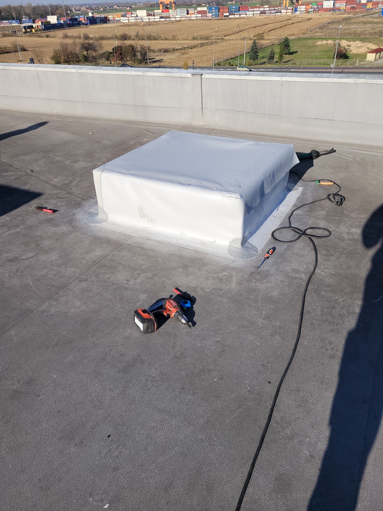 Szary dach płaski z obróbką komina w białej membranie, widoczny wiertarko-wkrętak akumulatorowy, opalarka dekarska i wkrętak płaski, w tle zabudowania przemysłowe