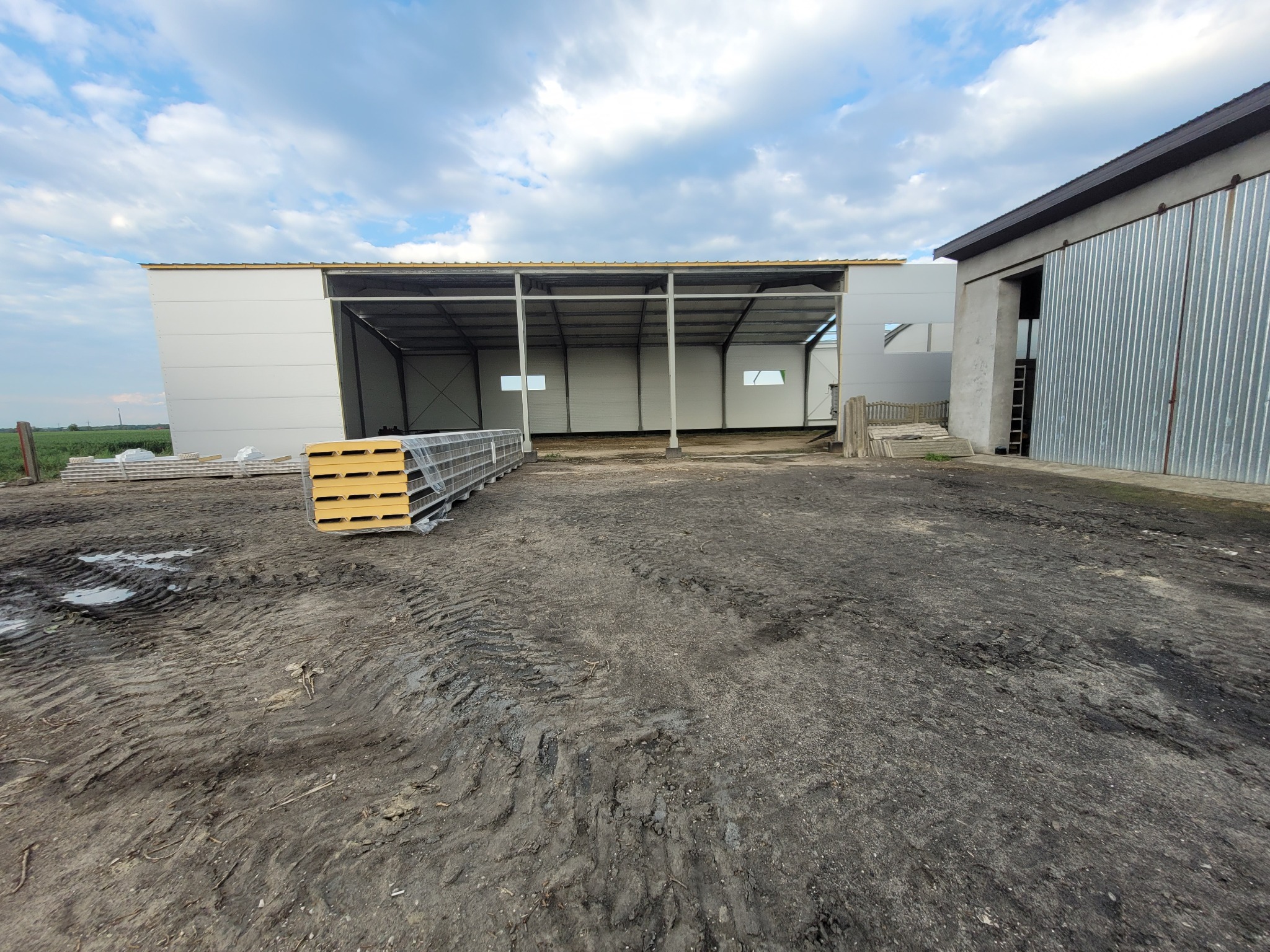 Budowa hali: widok na szkielet konstrukcji z zamontowanymi panelami ściennymi, w tle sterta paneli izolacyjnych i budynek z blachy falistej.