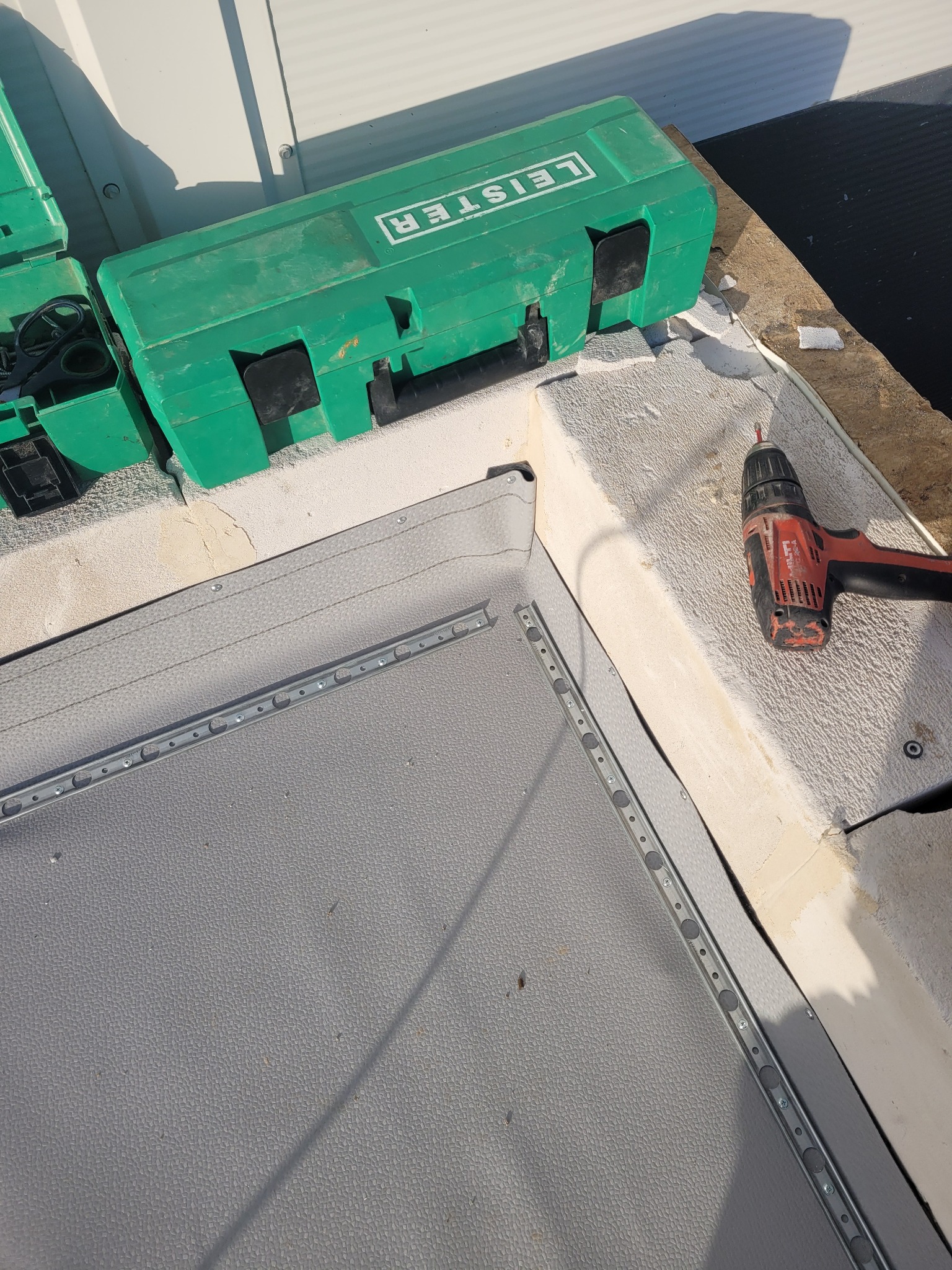 Szary dach z zamontowaną listwą montażową, zielona skrzynka narzędziowa Leister i wiertarko-wkrętarka Hilti obok krawędzi dachu.