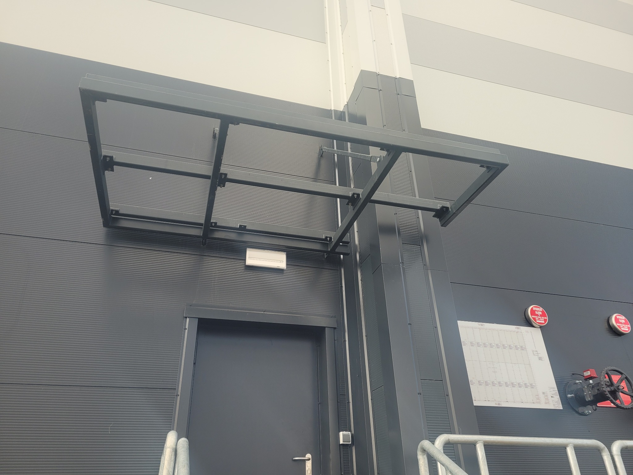 Szara metalowa konstrukcja daszku nad wejściem do budynku o panelowej elewacji, widoczne elementy instalacji przeciwpożarowej.