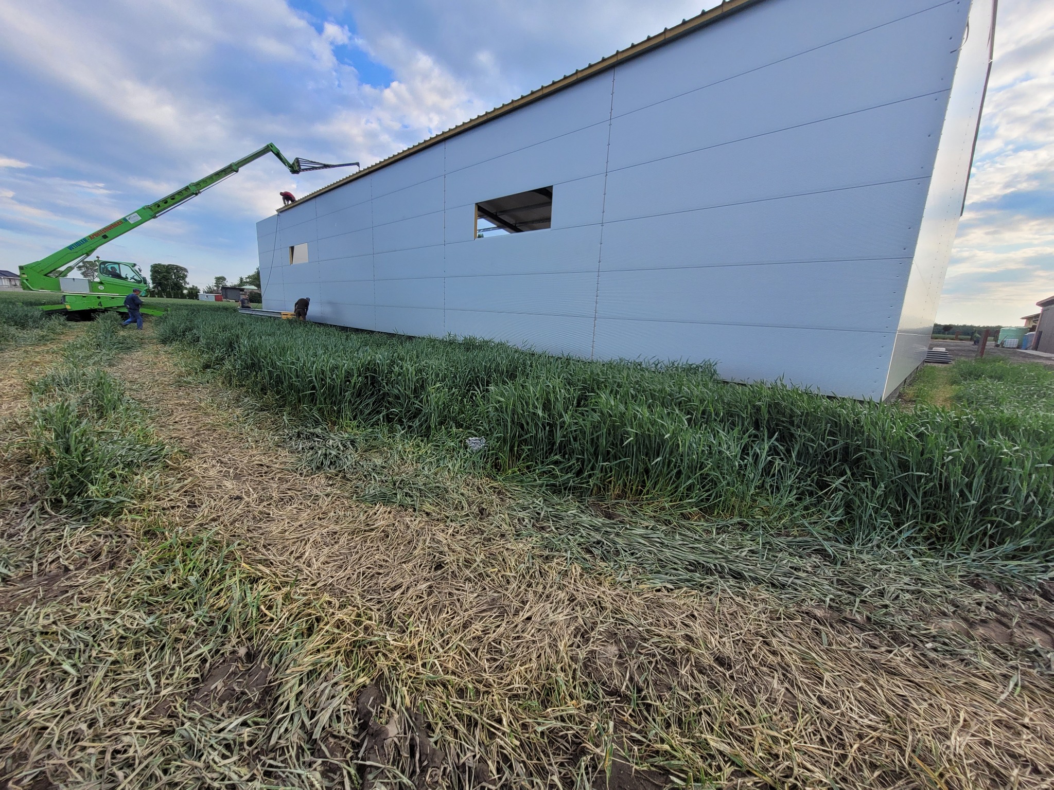 Montaż paneli ściennych na budynku przemysłowym, widoczny podnośnik koszowy i pracownicy na dachu, niezagospodarowany teren wokół budynku.