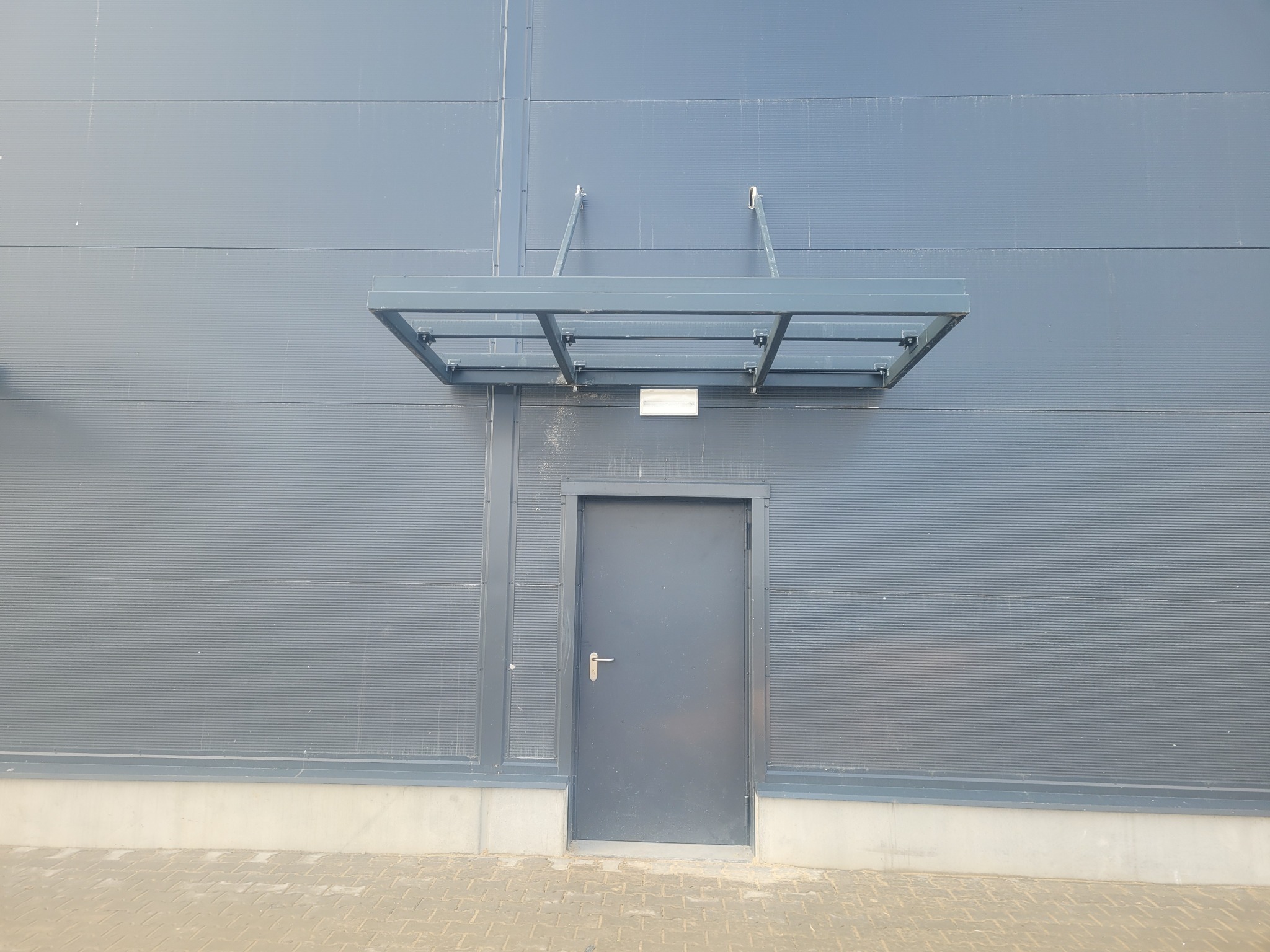 Szara, metalowa ściana budynku z zamontowanym daszkiem nad drzwiami wejściowymi, widoczne mocowania daszku.