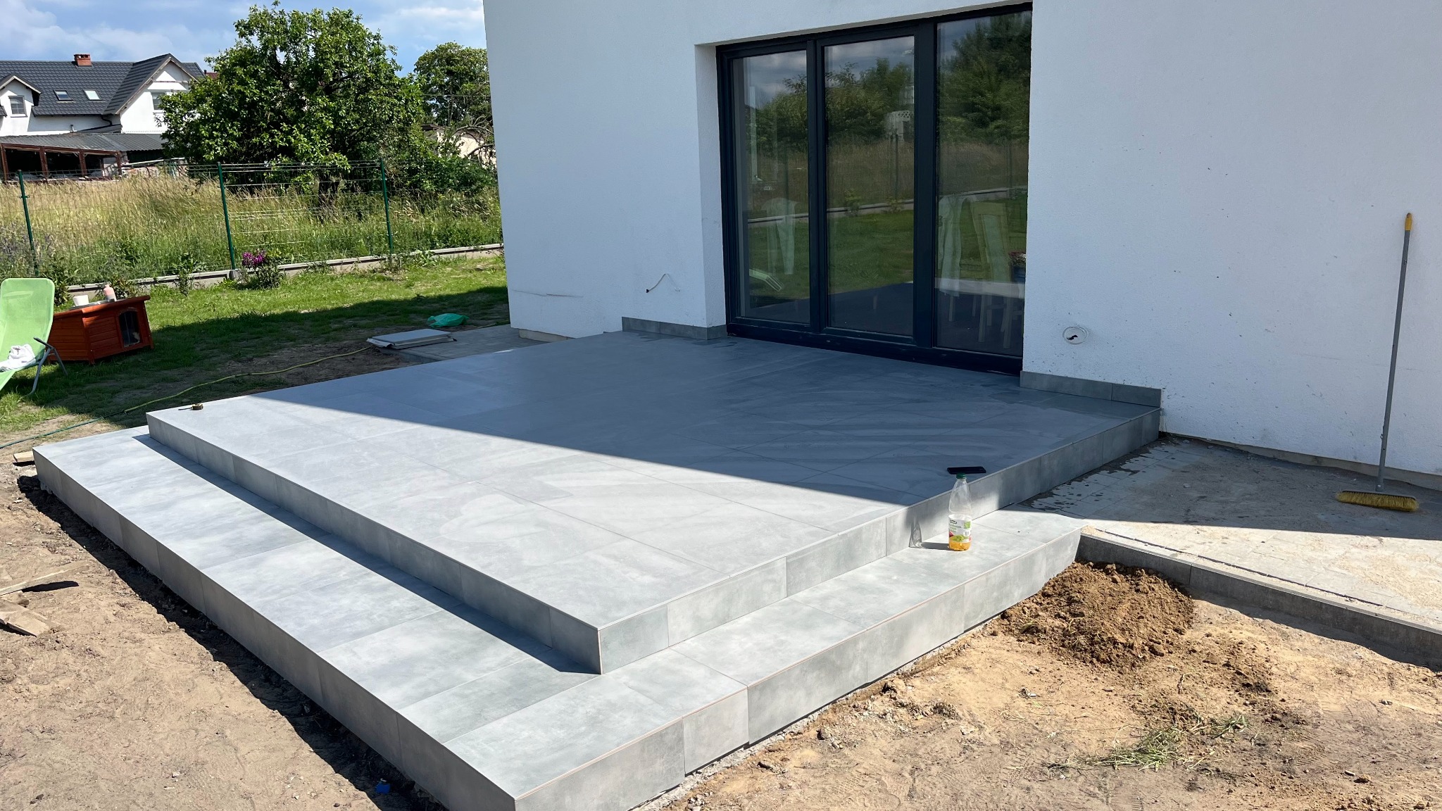 Wyłożony szarymi płytkami taras z trzema stopniami przed nowoczesnym domem z przesuwnymi drzwiami balkonowymi, widoczna sterta ziemi i butelka z płynem czyszczącym.