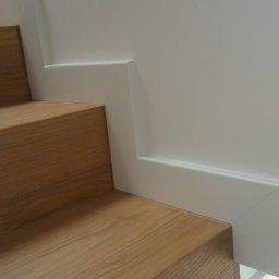 Z miłości do drewna Marcin Haber - Fragment schodów drewnianych z białą listwą przypodłogową przy ścianie, widok z dołu.