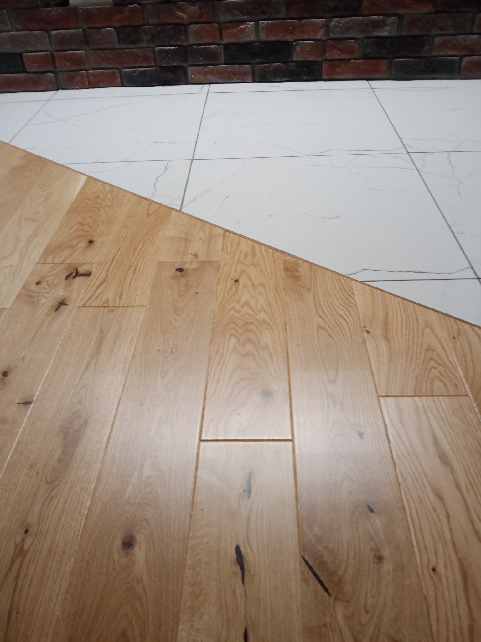 Połączenie jasnego parkietu z białymi płytkami imitującymi marmur i fragmentem ściany z cegły klinkierowej w tle.