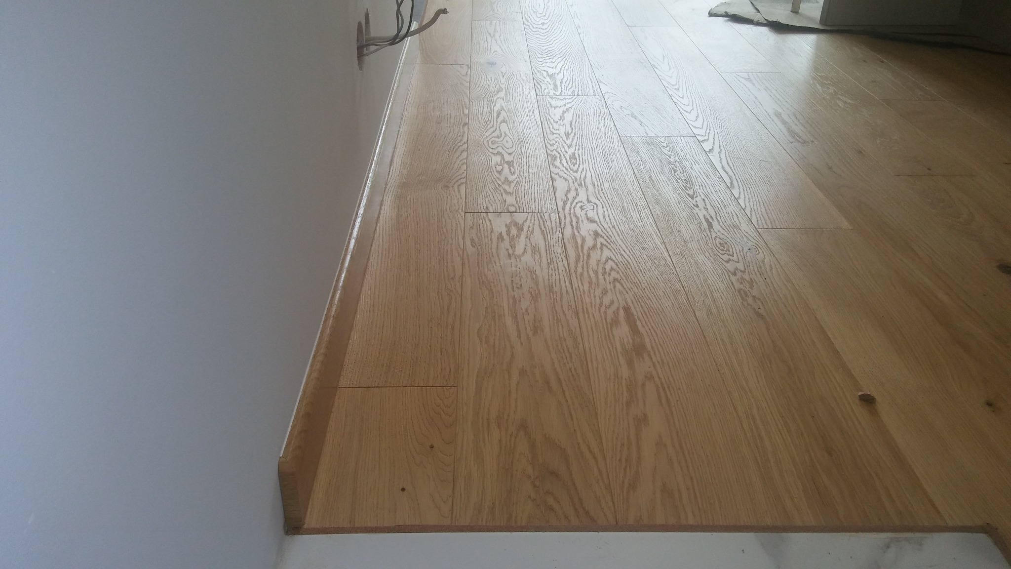 Jasny parkiet dębowy ułożony wzdłuż ściany, widoczne listwy przypodłogowe i wystające kable elektryczne z puszki w ścianie.