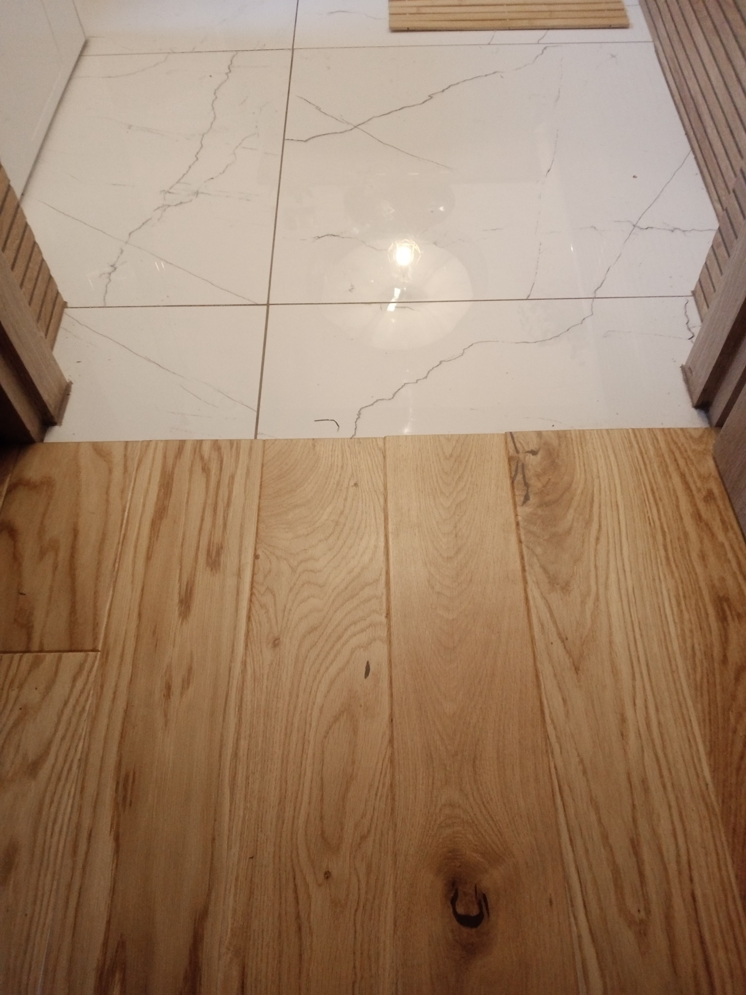 Parkiet z jasnego drewna styka się z białymi płytkami imitującymi marmur, widok z góry na podłogę przy drzwiach.