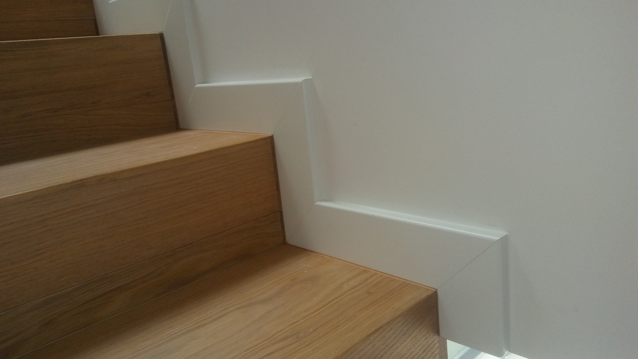 Fragment schodów drewnianych z białą listwą przypodłogową przy ścianie, widok z dołu.
