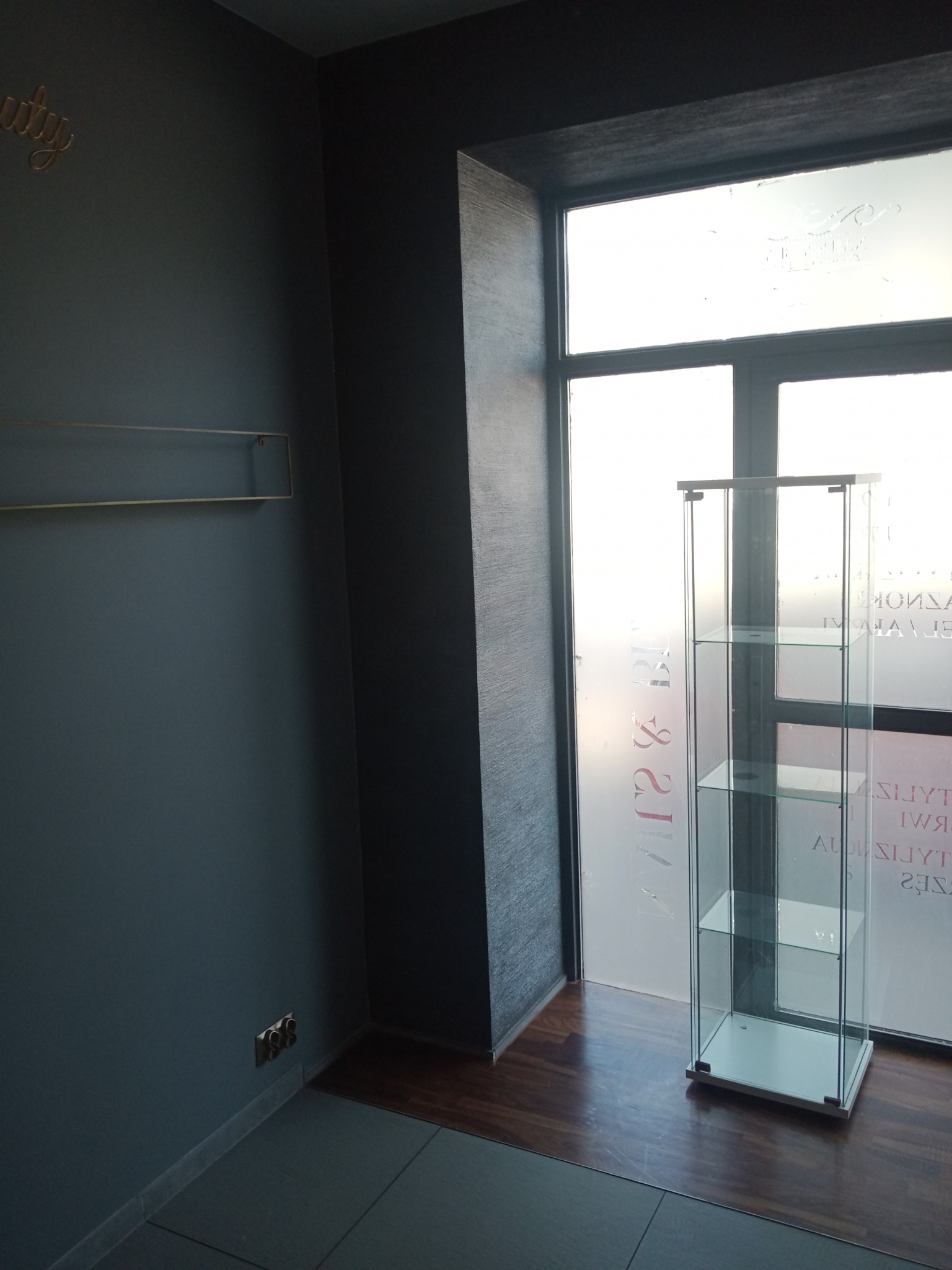 Pomalowane na szaro pomieszczenie z przeszkloną witryną na tle okna z naklejonymi napisami, widoczne gniazdko elektryczne przy podłodze oraz dekoracja ścienna.