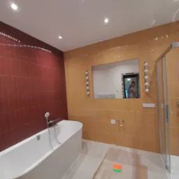 Nowoczesna łazienka z dwukolorowymi płytkami, białą wanną i lustrem, w którym odbija się osoba robiąca zdjęcie. Podłoga zabezpieczona kartonem i folią malarską.