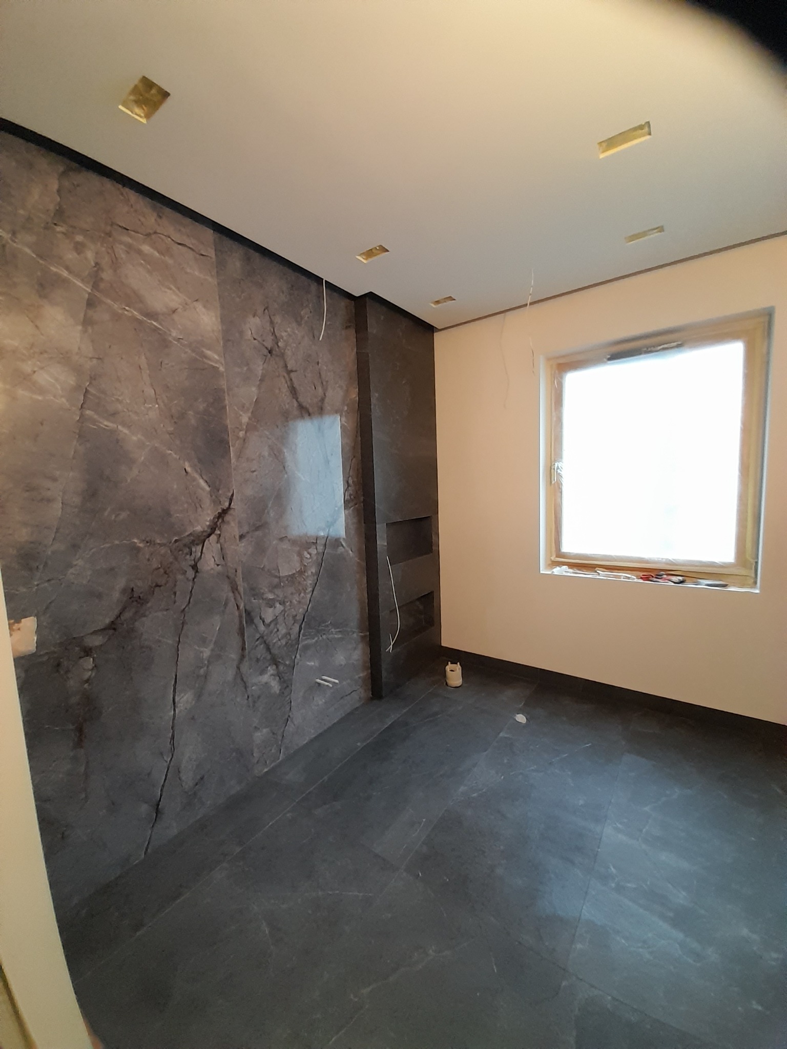 Wnętrze pokoju z wykończoną ścianą imitującą marmur, czarną podłogą z płytek, oknem zabezpieczonym folią i przygotowanymi otworami na oświetlenie w suficie.