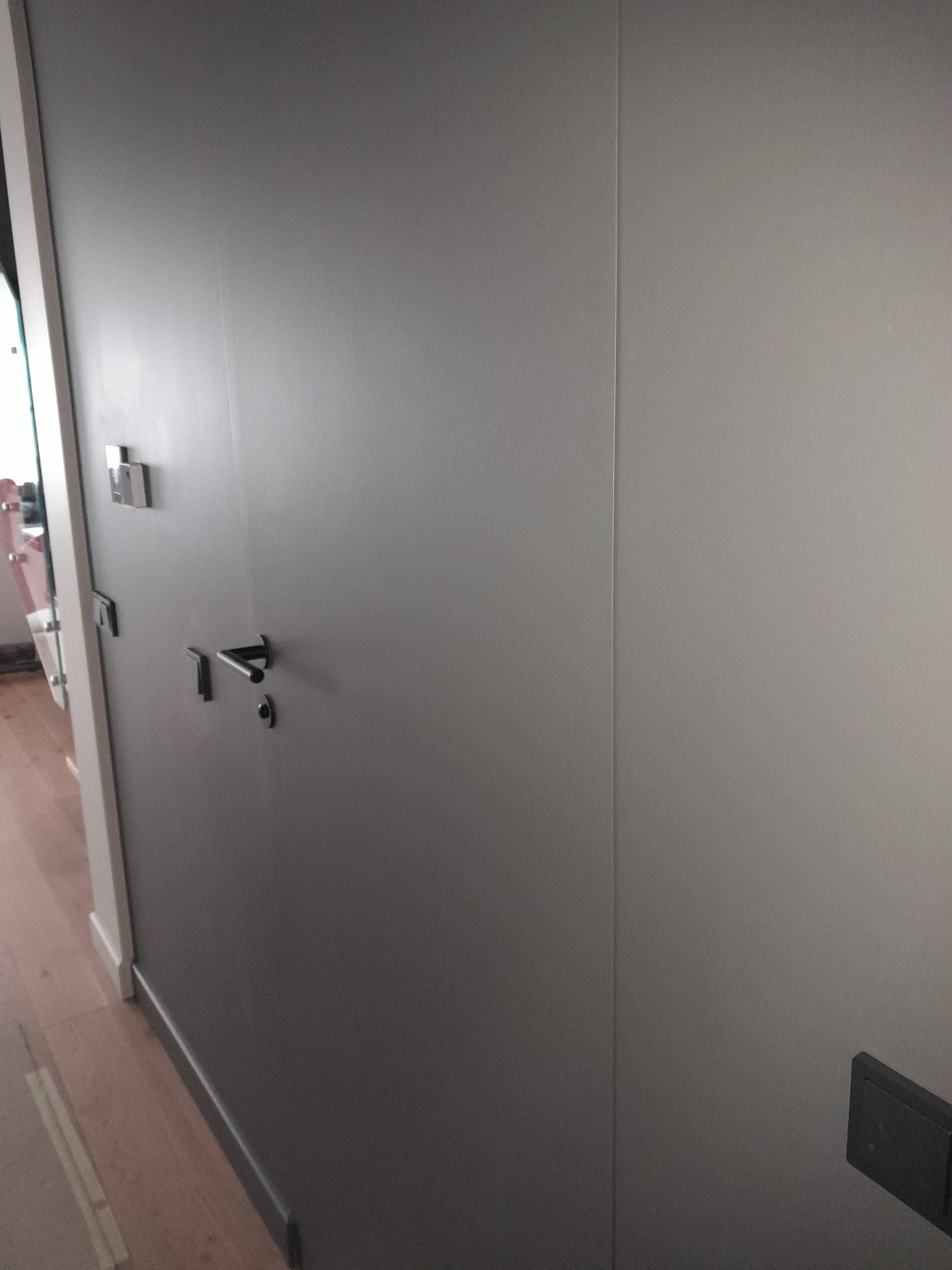 Ściana w odcieniach szarości z zamontowaną klamką i włącznikiem światła, widoczna linia łączenia płyt. Podłoga wykończona panelami.