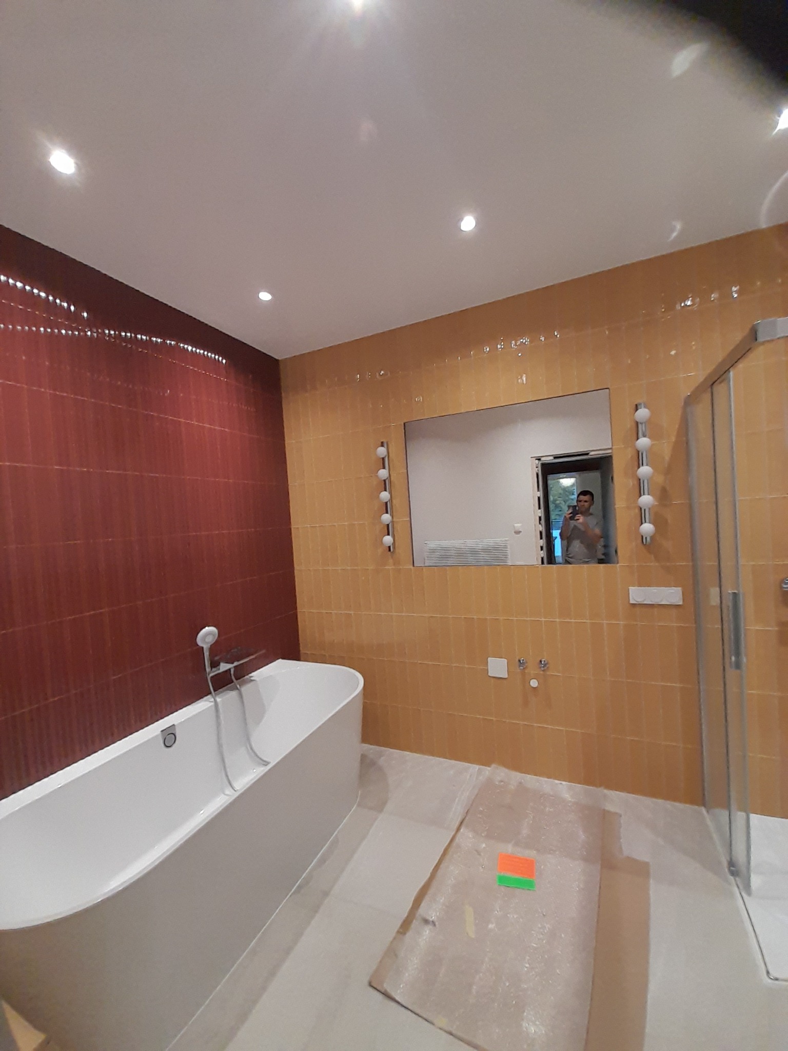 Nowoczesna łazienka z dwukolorowymi płytkami, białą wanną i lustrem, w którym odbija się osoba robiąca zdjęcie. Podłoga zabezpieczona kartonem i folią malarską.