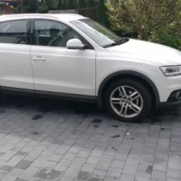 Biały samochód osobowy, Audi Q3, zaparkowany na kostce brukowej, widok z boku, na tle zieleni ogrodowej. Auto czyste, zadbane, felgi aluminiowe.