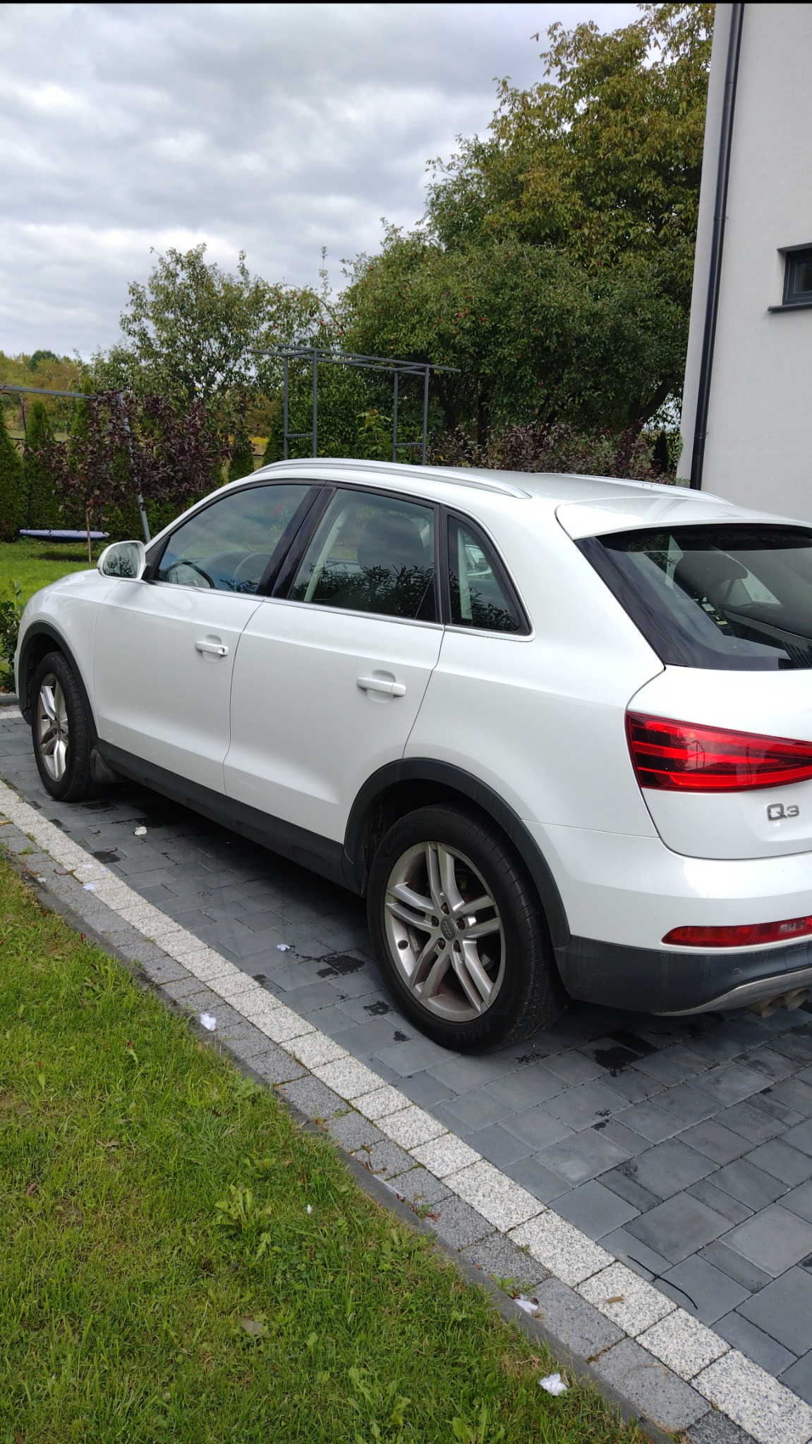 Biały SUV Audi Q3 zaparkowany na posesji z kostki brukowej, widok z tyłu i boku, na tle zieleni i białego budynku. Model Q3 widoczny na tylnej lampie.