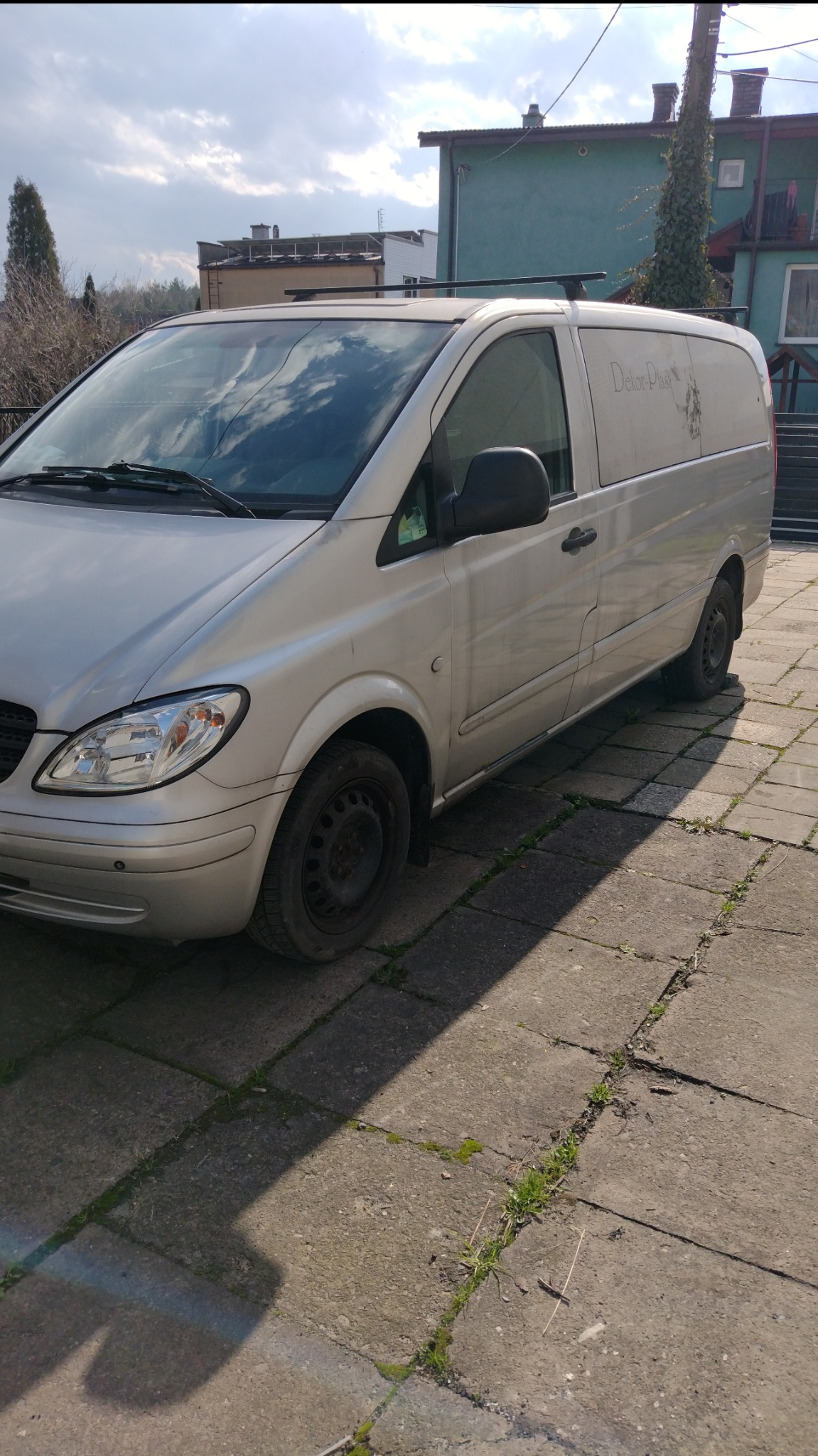 Srebrny bus marki Mercedes-Benz Vito z napisem 'Dekor-Plast' na boku, zaparkowany na brukowanym podjeździe przed budynkiem w słoneczny dzień.