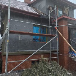 DOMLUX DOROTA ROMANOWSKA - Ocieplanie elewacji domu jednorodzinnego styropianem grafitowym, widoczne rusztowania i pracownicy w trakcie montażu, okna zabezpieczone folią.