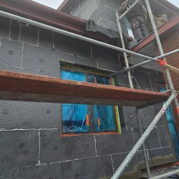 DOMLUX DOROTA ROMANOWSKA - Ocieplanie budynku styropianem grafitowym, zabezpieczone okna folią, widoczne rusztowanie i pracownicy na wysokości.