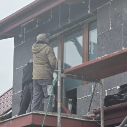 DOMLUX DOROTA ROMANOWSKA - Dwóch pracowników na rusztowaniu montuje okno w ścianie z betonu komórkowego z widocznymi śladami pianki montażowej.