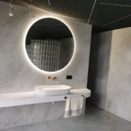 Nowoczesna łazienka z szarymi płytkami imitującymi marmur, okrągłym lustrem z podświetleniem LED, białą umywalką nablatową na długim blacie i czarnym sufitem z białymi listwami.