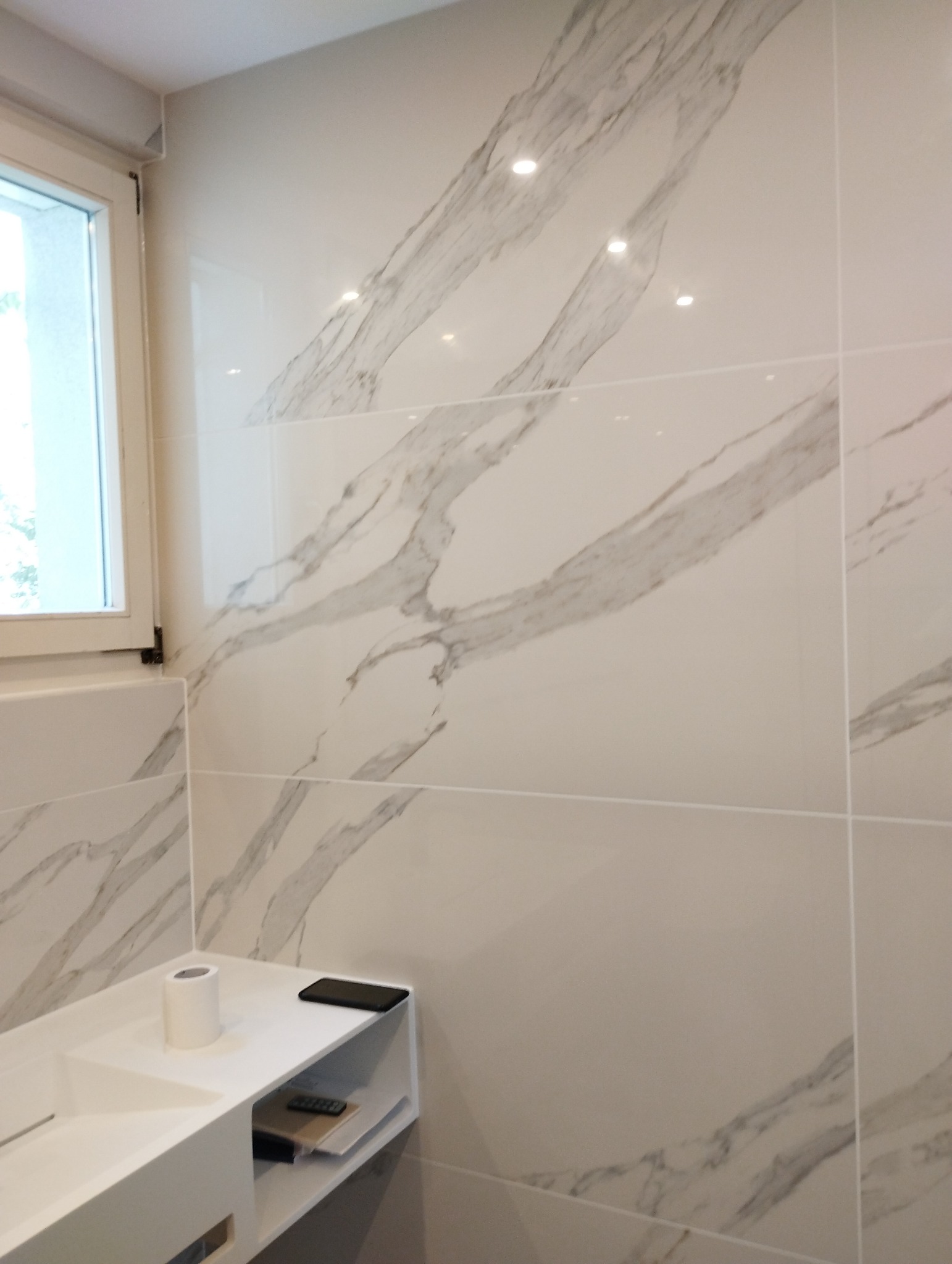 Ściana łazienki wyłożona dużymi, białymi płytkami z szarym marmurowym wzorem, z umywalką i toaletą w tle, widok z bliska.