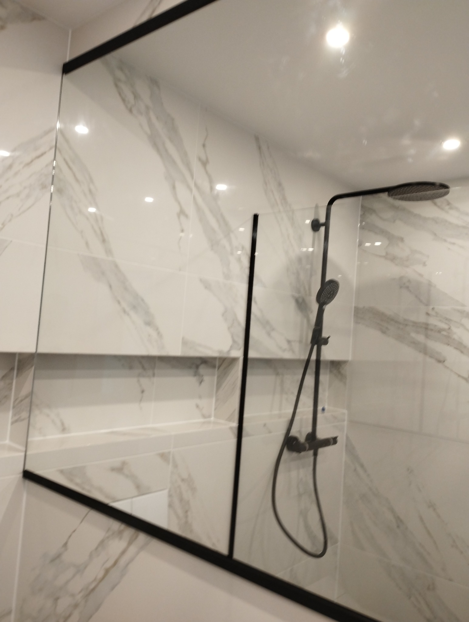 Nowoczesna kabina prysznicowa typu walk-in z czarnymi profilami, marmurowe płytki na ścianach, deszczownica i słuchawka prysznicowa w kolorze czarnym.