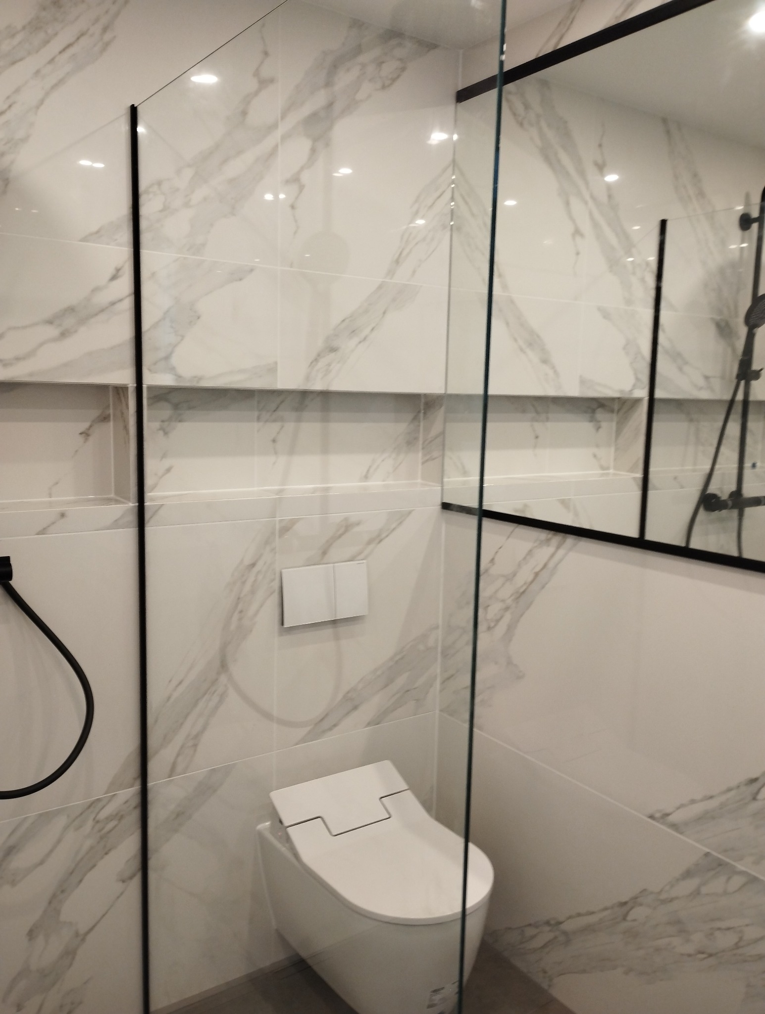 Nowoczesna łazienka z białą armaturą, marmurowe płytki na ścianach, czarne ramy kabiny prysznicowej i wnęka w ścianie.