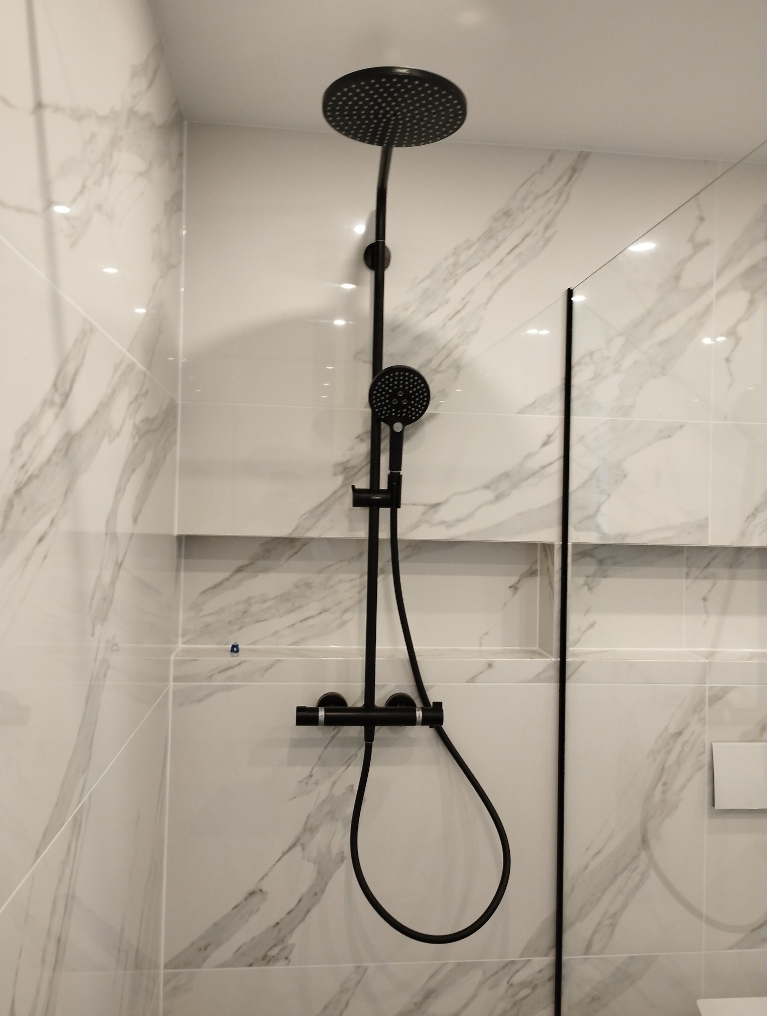 Czarny zestaw natryskowy z deszczownicą i słuchawką, zamontowany na ścianie z białych płytek imitujących marmur w nowoczesnej łazience.