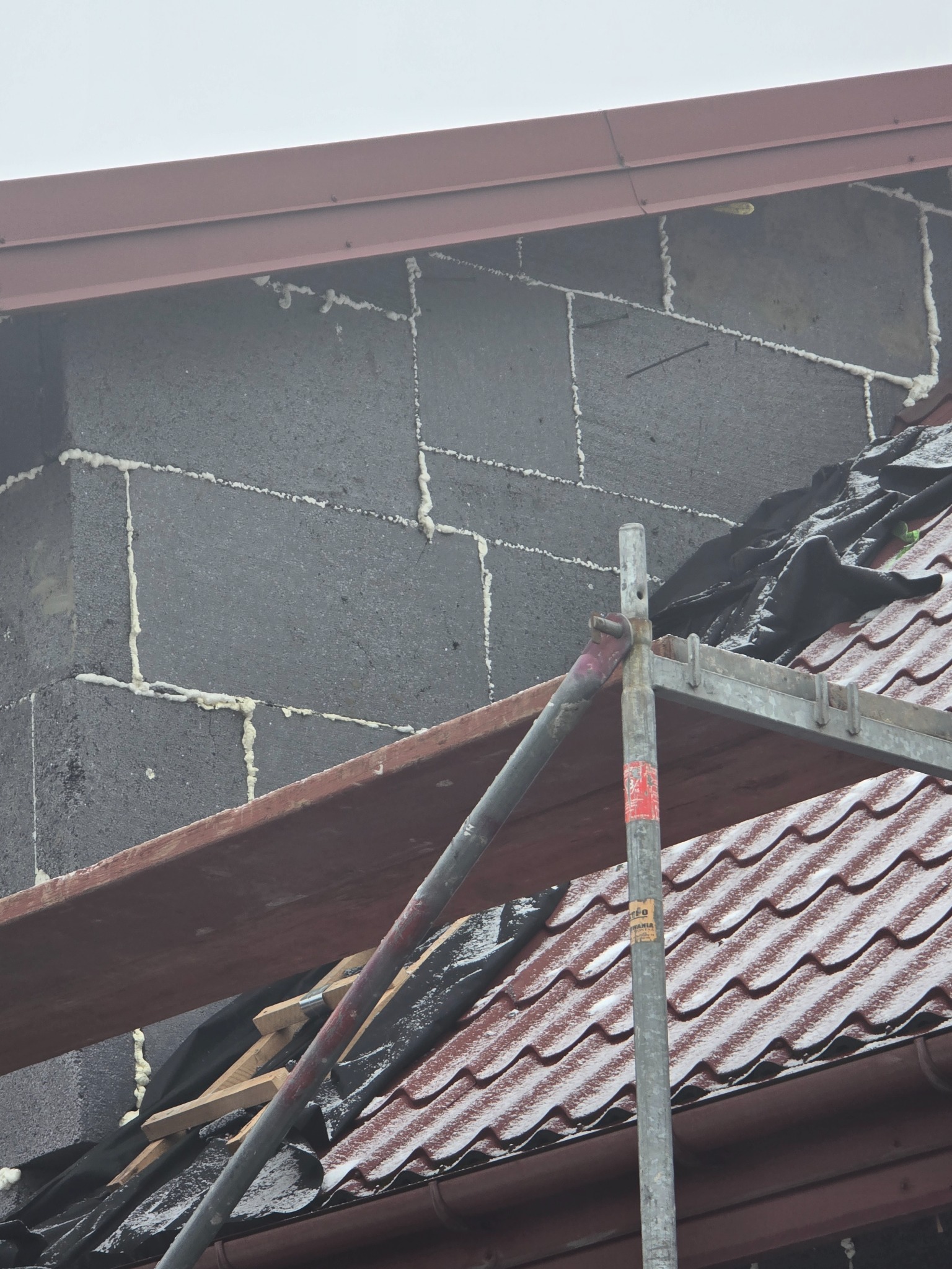 Ocieplony komin z szarego bloczka z widocznymi spoinami wypełnionymi pianką montażową, zabezpieczony rusztowaniem na tle dachu pokrytego czerwoną dachówką.