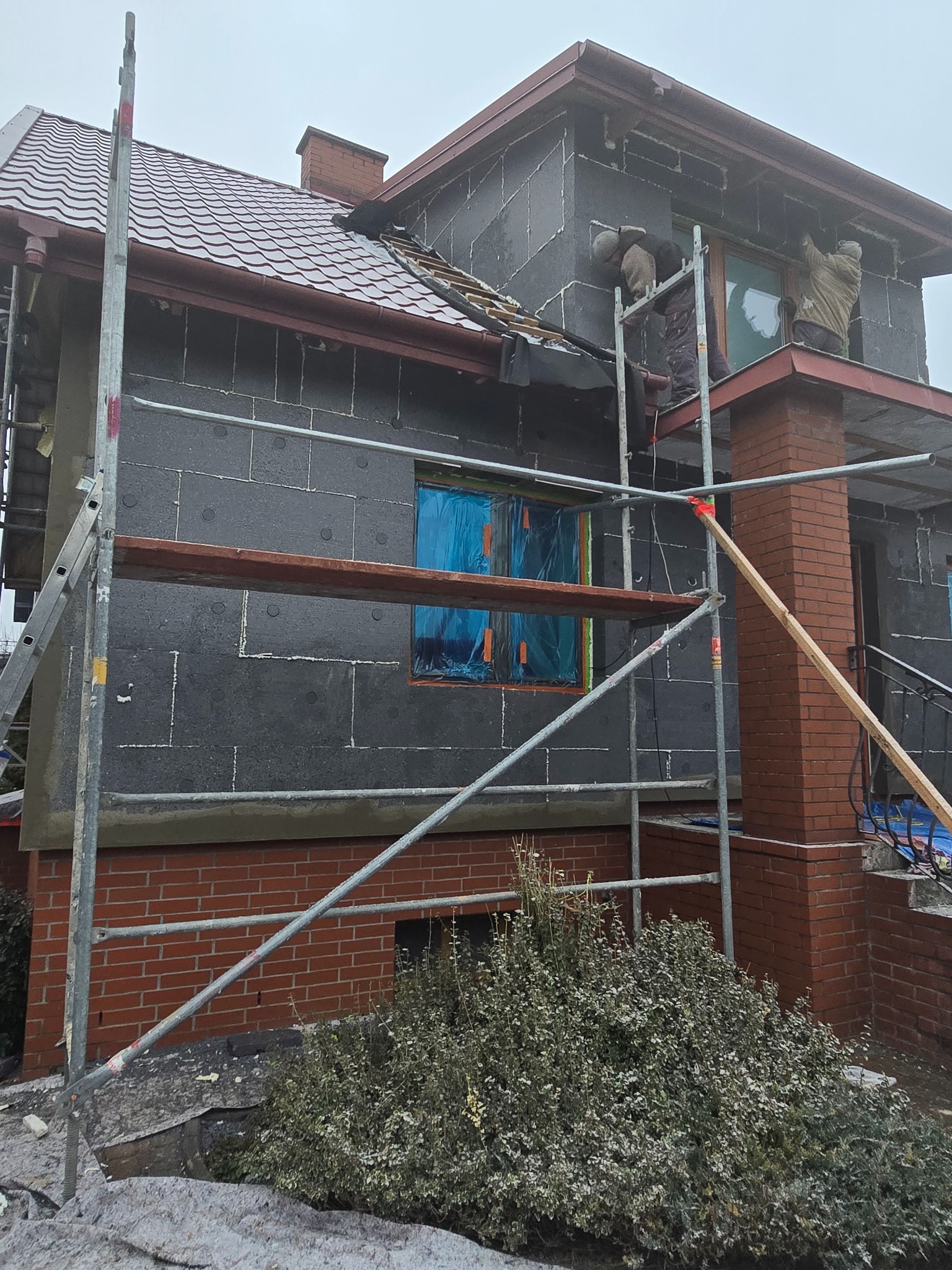 Ocieplanie elewacji domu jednorodzinnego styropianem grafitowym, widoczne rusztowania i pracownicy w trakcie montażu, okna zabezpieczone folią.