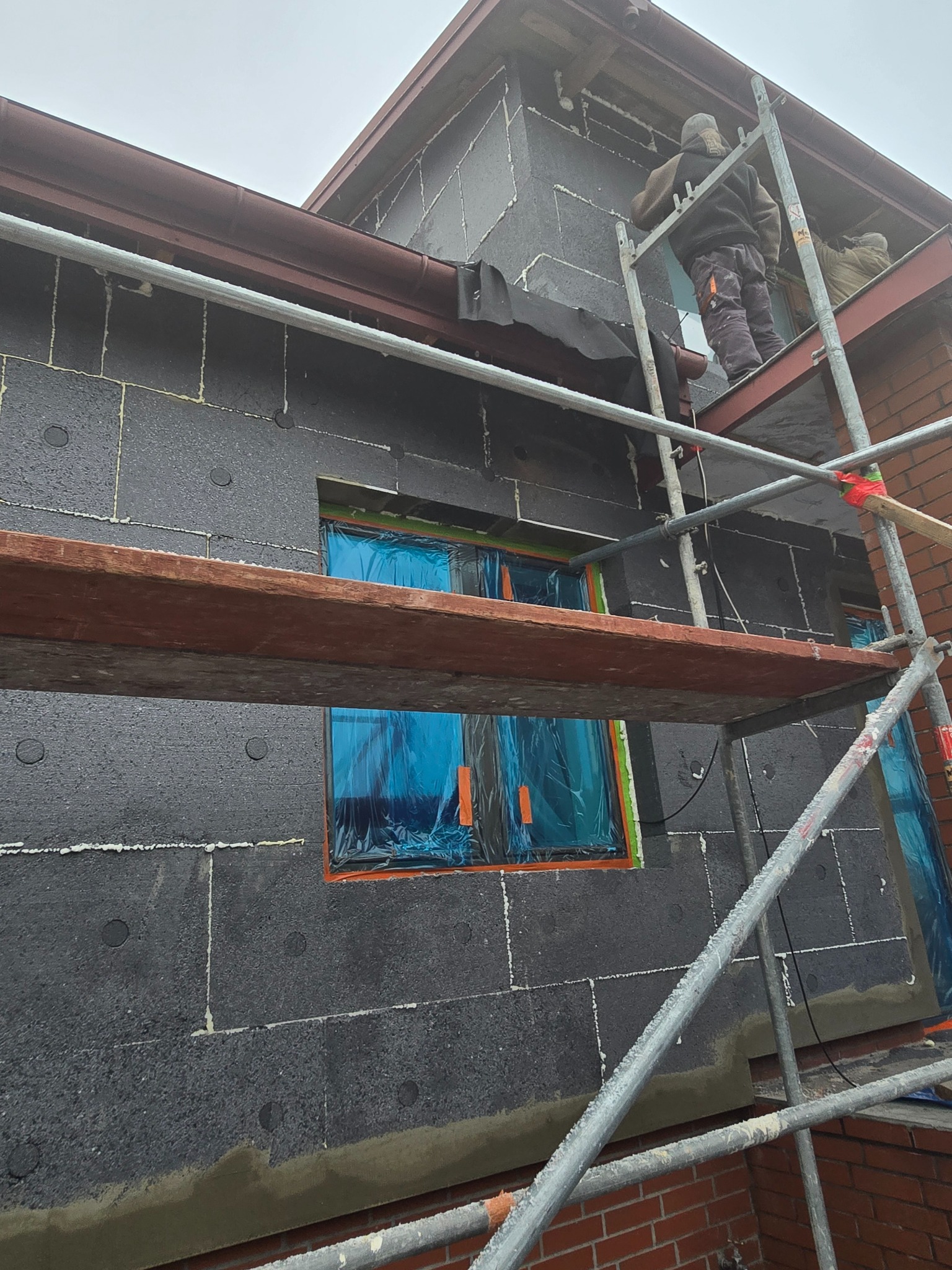 Ocieplanie budynku styropianem grafitowym, zabezpieczone okna folią, widoczne rusztowanie i pracownicy na wysokości.