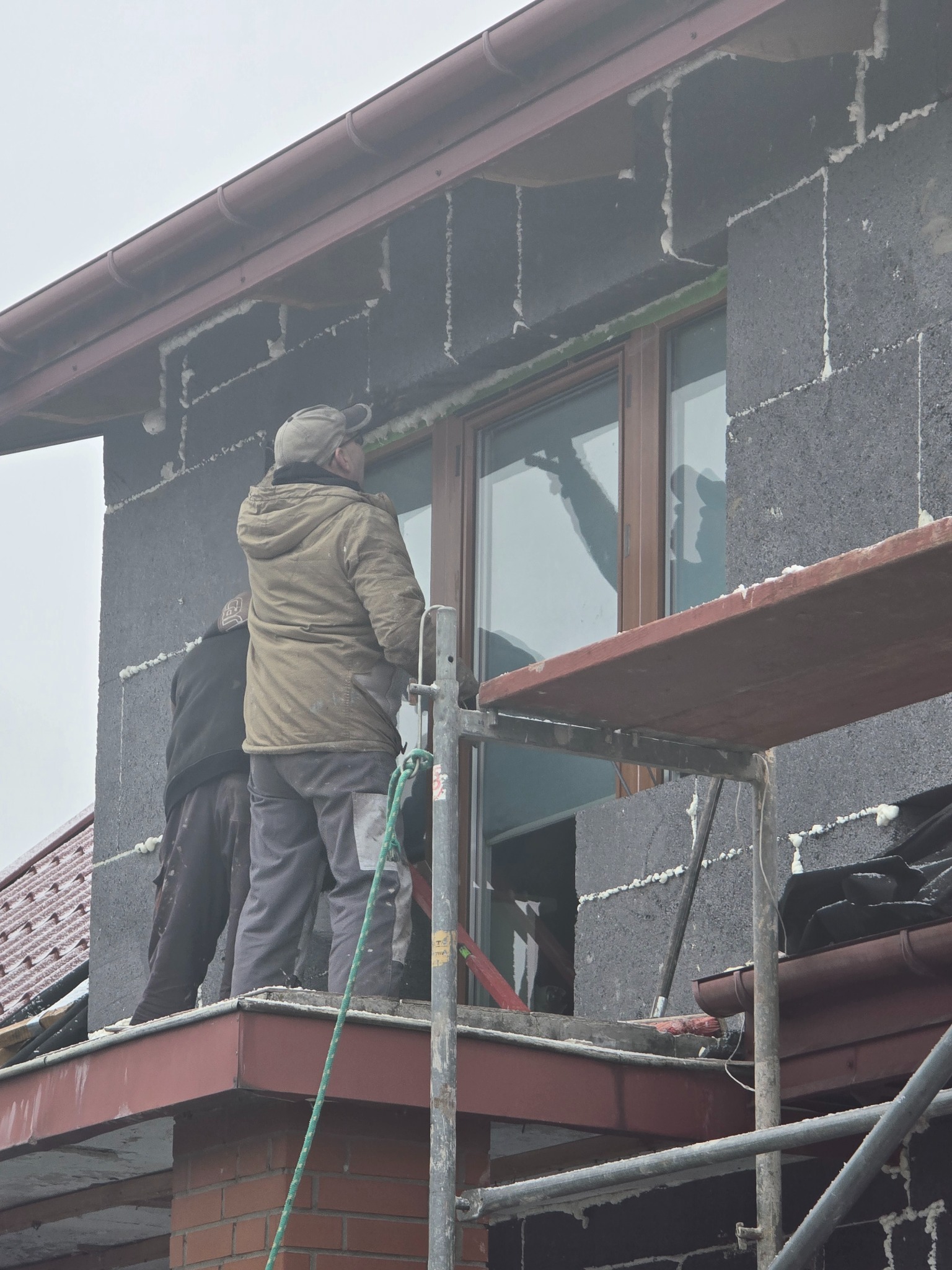 Dwóch pracowników na rusztowaniu montuje okno w ścianie z betonu komórkowego z widocznymi śladami pianki montażowej.