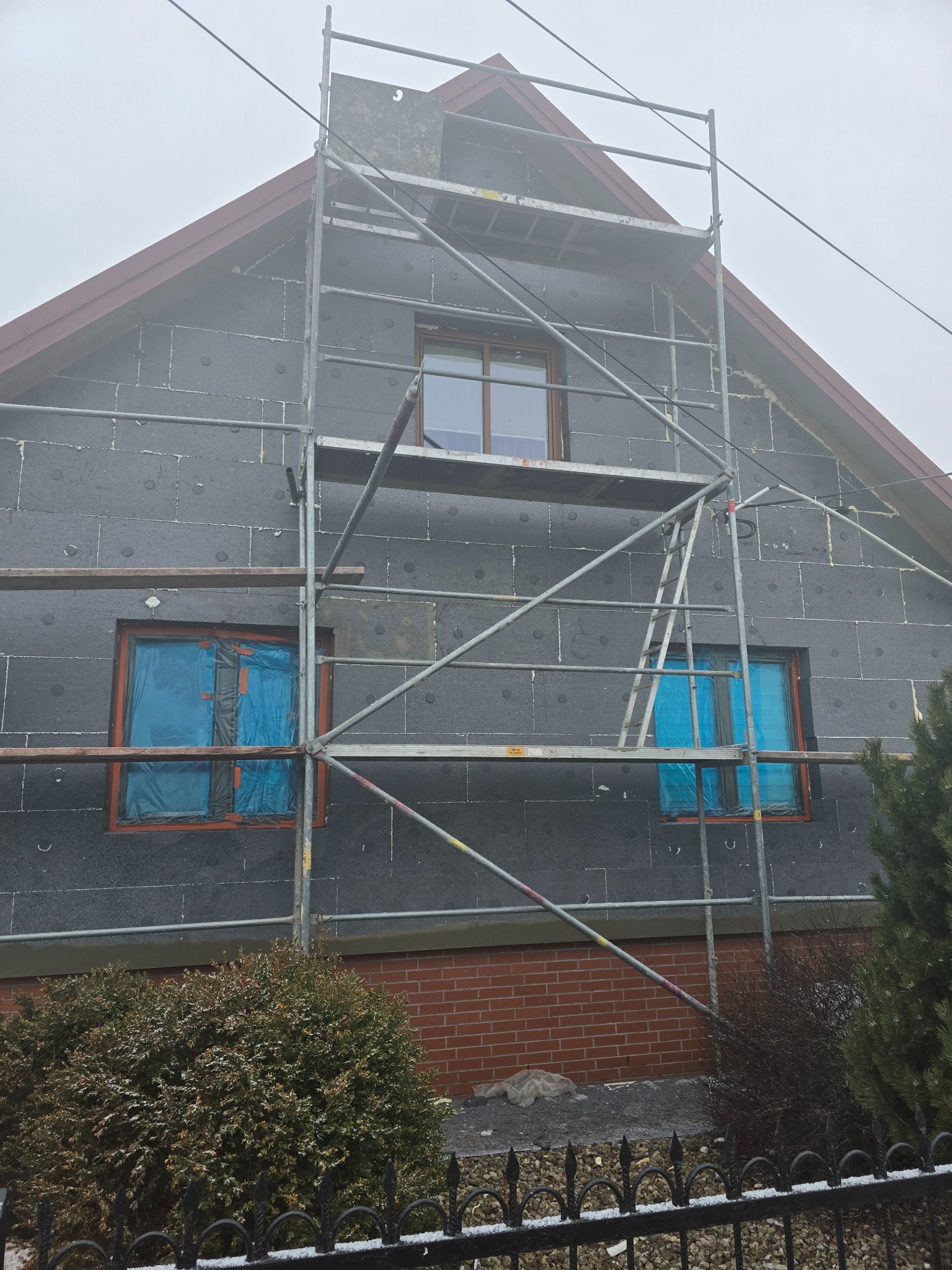 Dom jednorodzinny w trakcie ocieplania styropianem grafitowym, widoczne rusztowanie i okna zabezpieczone folią, pochmurna pogoda.
