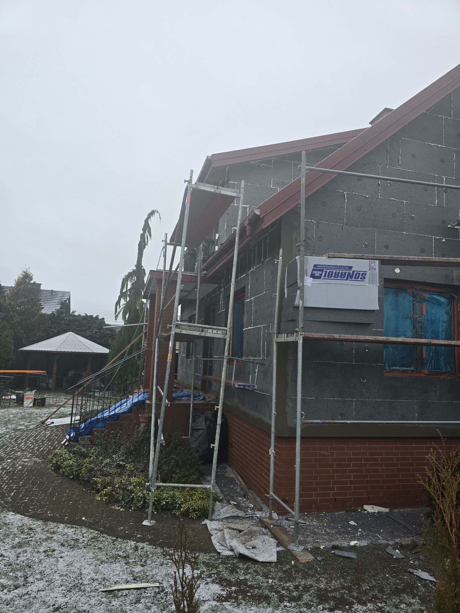 Ocieplanie elewacji domu jednorodzinnego czarnym styropianem, widoczne rusztowanie, okna zabezpieczone folią, opakowanie styropianu SONAROL