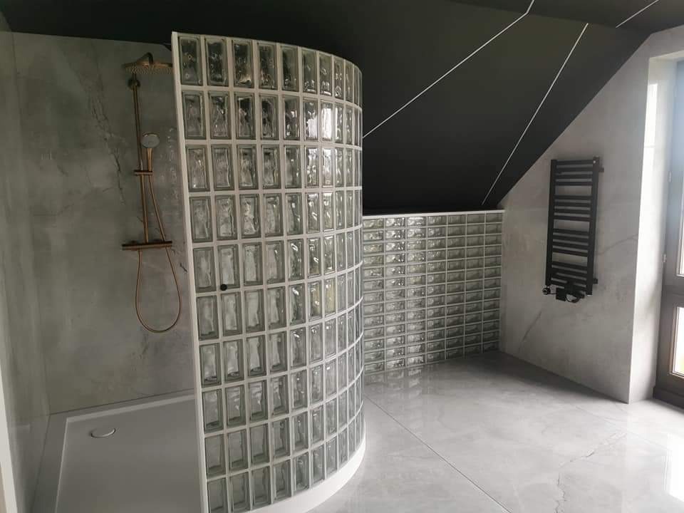 Nowoczesna łazienka na poddaszu z kabiną prysznicową oddzieloną ścianką z luksferów, szara armatura i płyty imitujące beton, czarny sufit z białymi liniami.