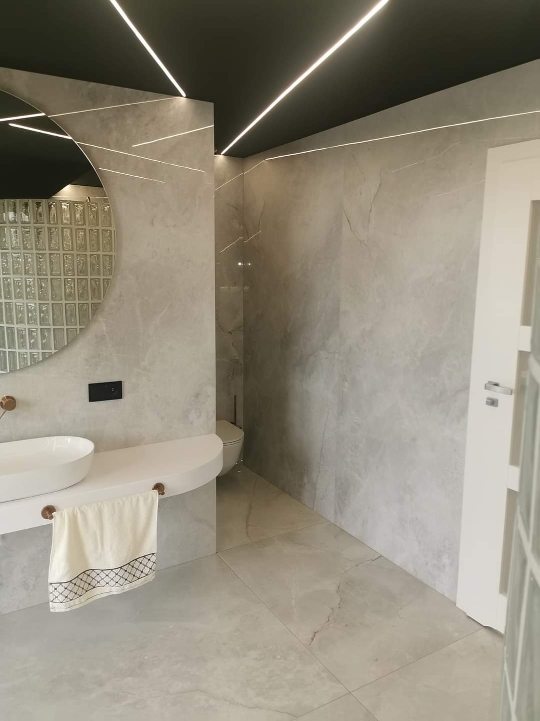 Nowoczesna łazienka z szarymi płytkami imitującymi marmur, okrągłym lustrem z ramą z luksferów, podwieszaną szafką z umywalką i oświetleniem LED na suficie.