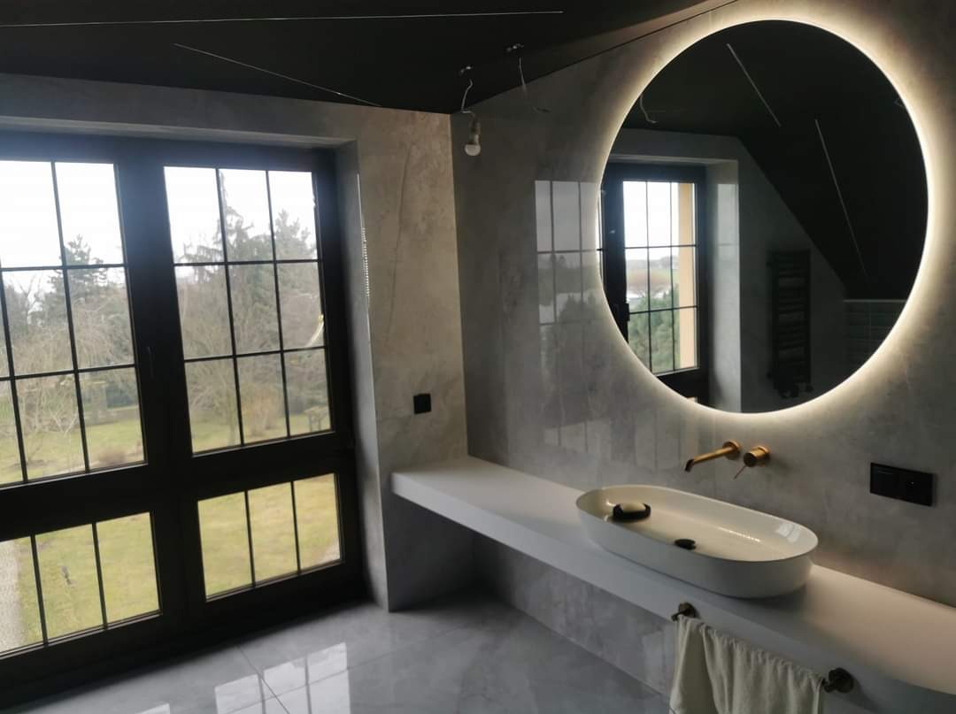 Nowoczesna łazienka z dużym oknem i okrągłym lustrem podświetlanym LED. Umywalka nablatowa o owalnym kształcie, bateria w kolorze szczotkowanego złota. Ściany wykończone w szarym dekoracyjnym tynku.