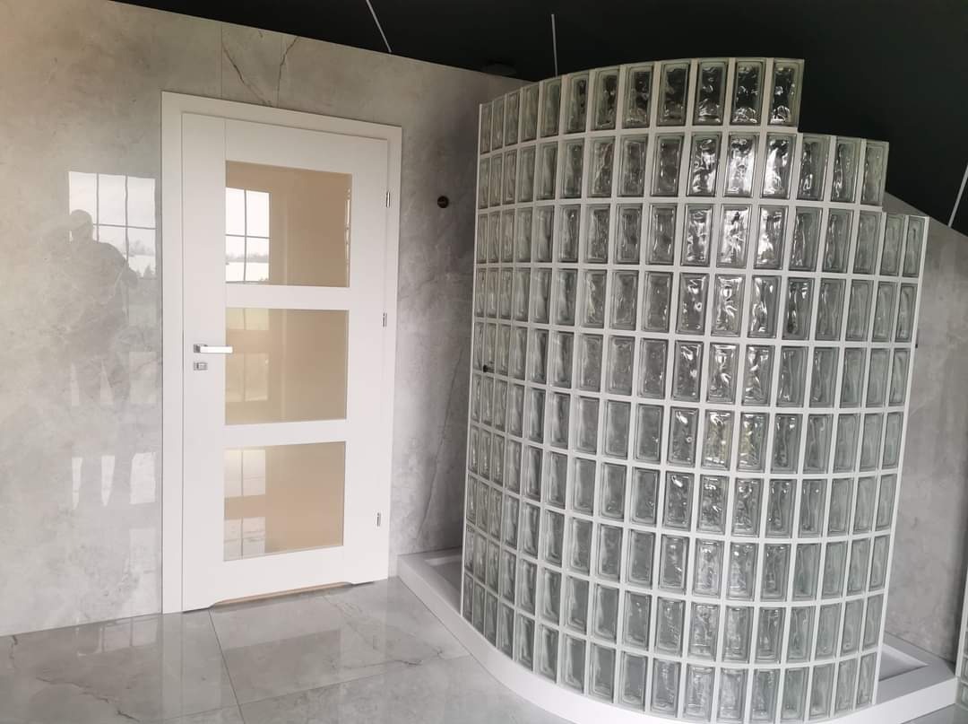 Białe drzwi z trzema szybami obok zaokrąglonej ścianki prysznicowej z luksferów na tle marmurowej ściany i podłogi.