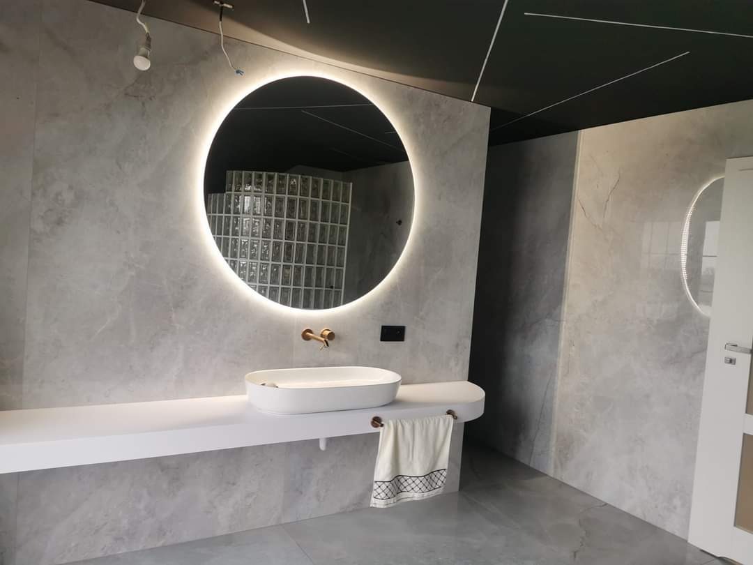 Nowoczesna łazienka z szarymi płytkami imitującymi marmur, okrągłym lustrem z podświetleniem LED, białą umywalką nablatową na długim blacie i czarnym sufitem z białymi listwami.