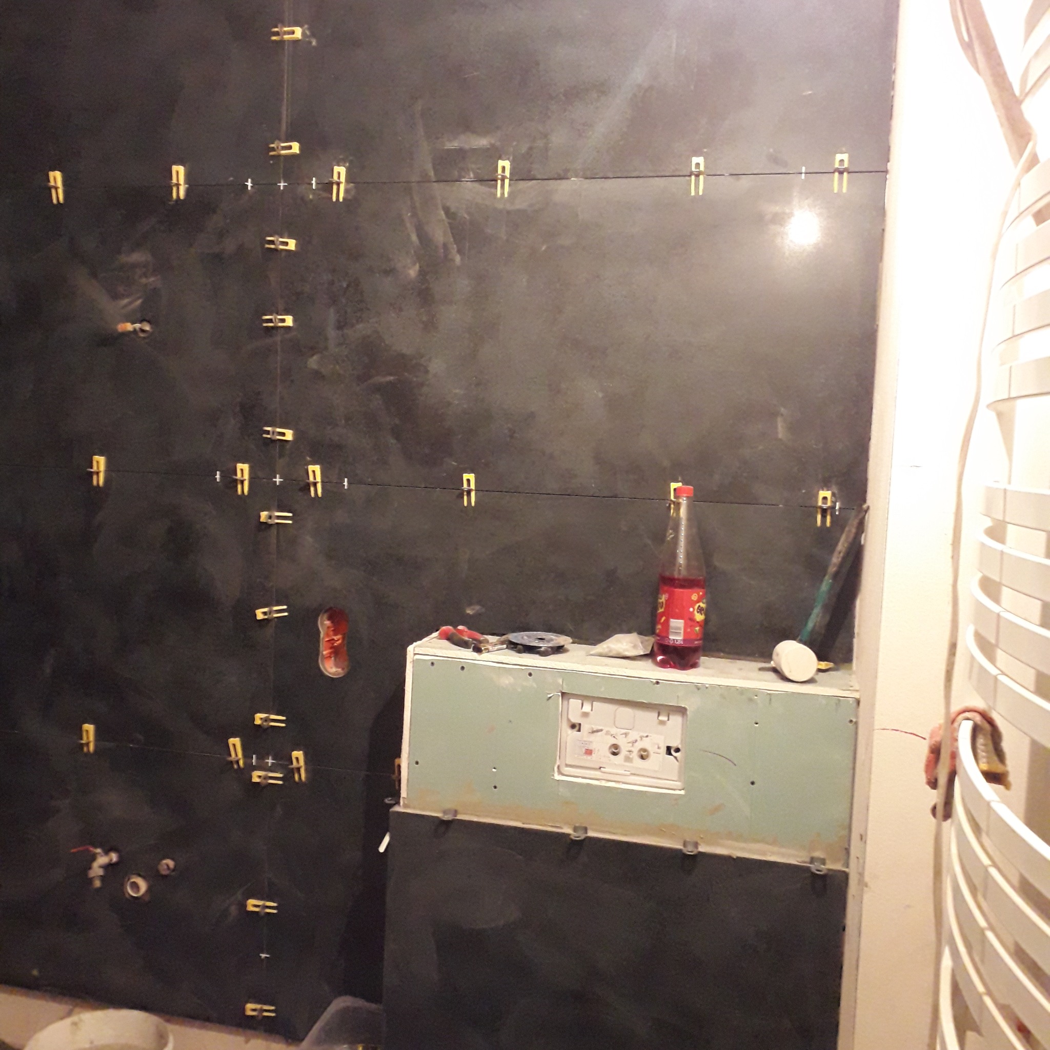 Ciemne płytki imitujące kamień, ułożone w łazience z użyciem systemu poziomowania płytek, widoczne narzędzia i butelka napoju na obudowie stelaża podtynkowego.