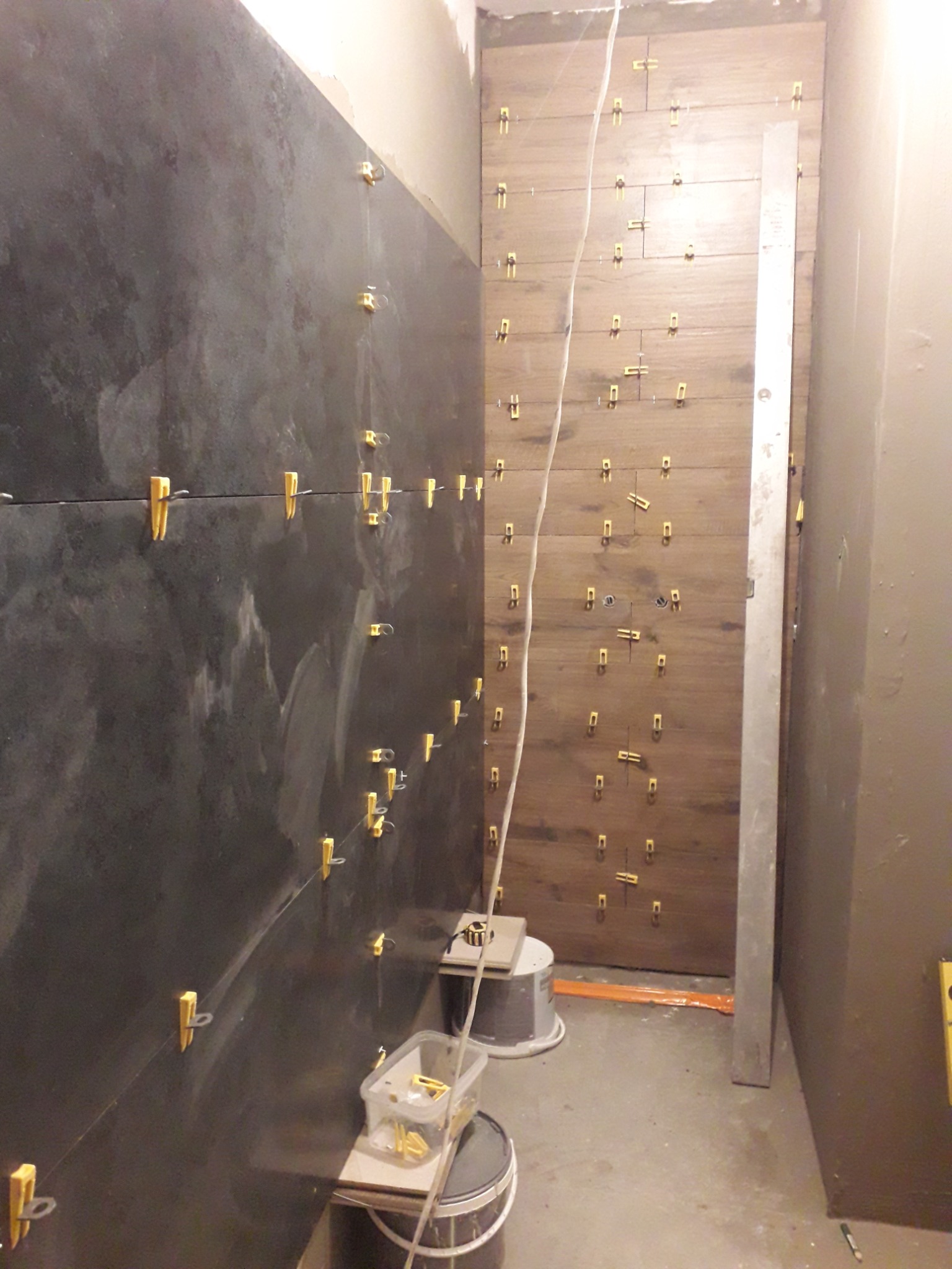 Układanie płytek w łazience: ciemne płytki na ścianie z żółtymi klipsami poziomującymi, obok ściana z jasnych płytek imitujących drewno, również z klipsami, widoczne wiaderka i poziomica.