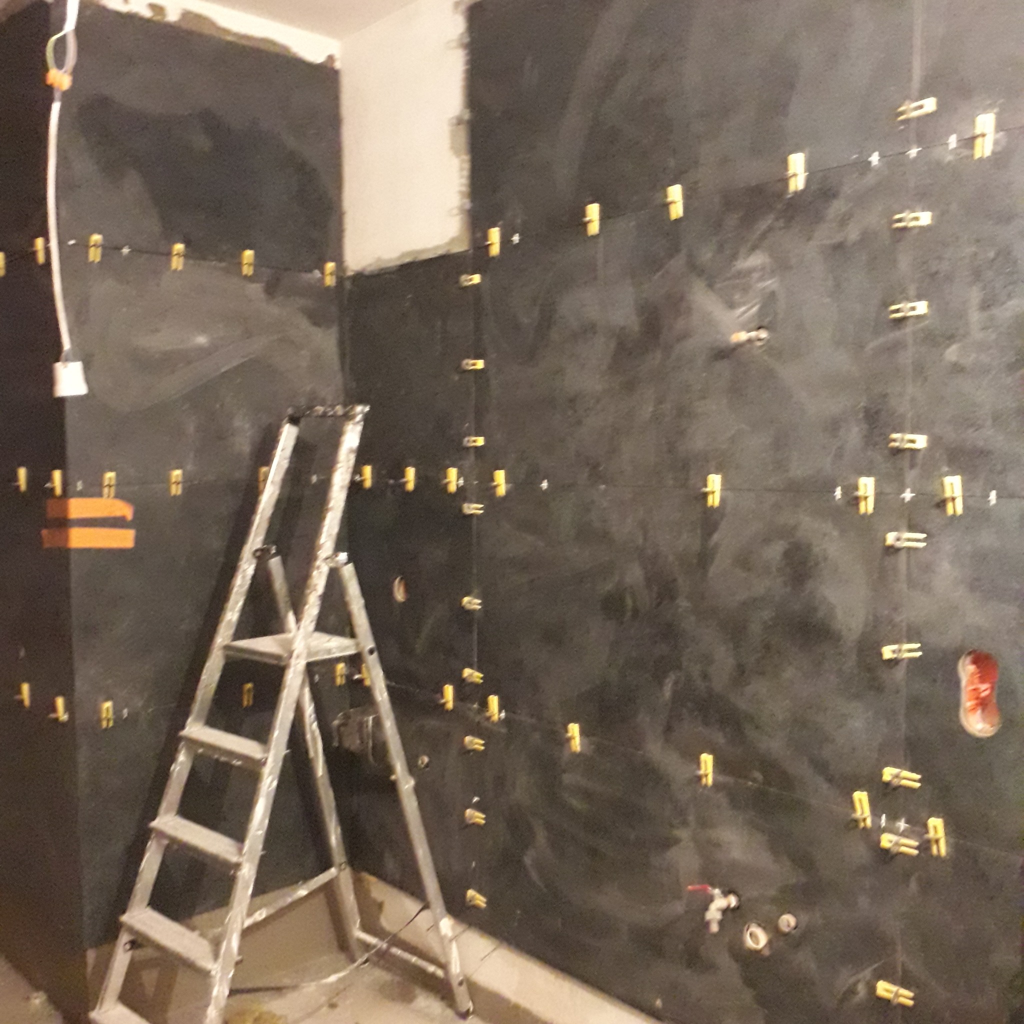 Ciemna, nierówna ściana w trakcie układania płytek, widoczne plastikowe krzyżyki dystansowe i poziomica laserowa, drabina aluminiowa oparta o ścianę.