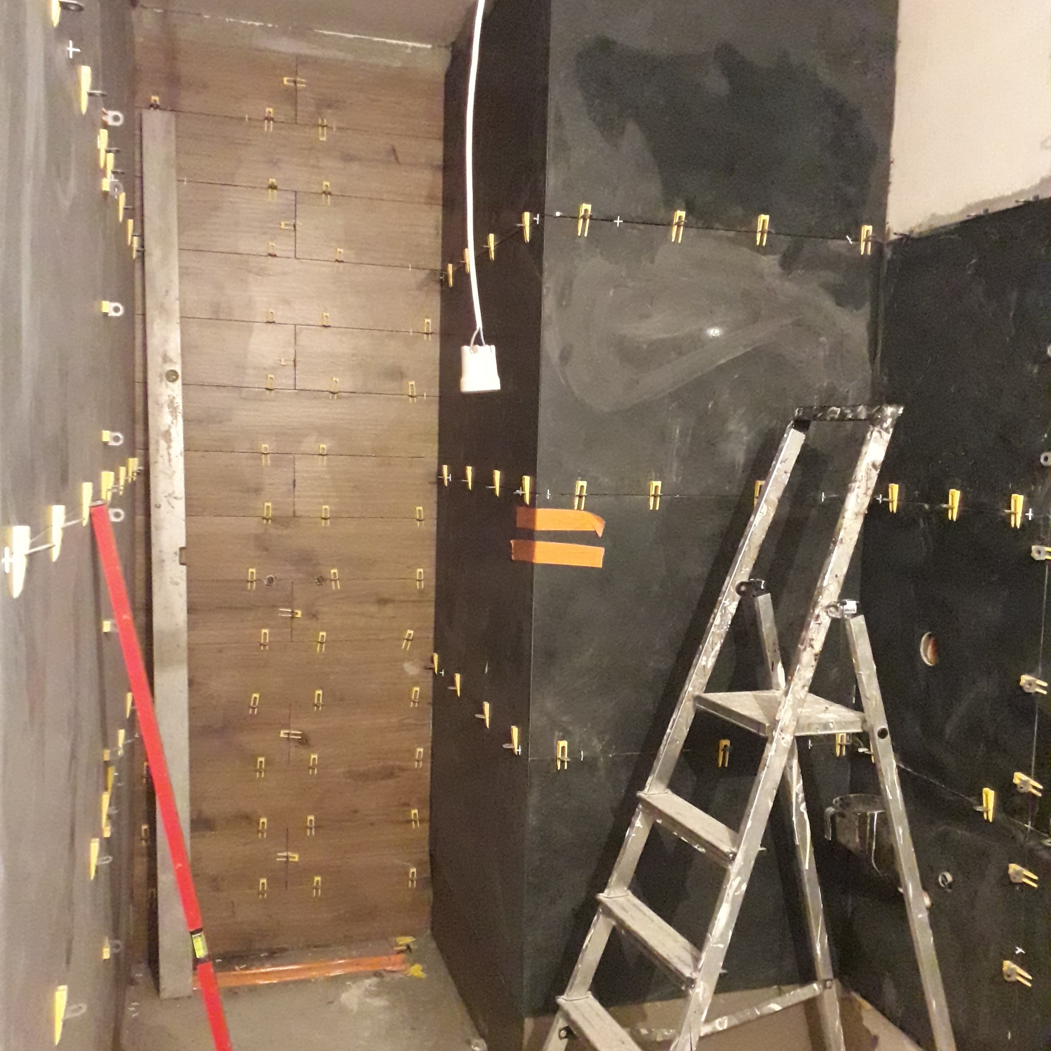 Świeżo ułożone płytki imitujące drewno i ciemne płytki na ścianach łazienki, z użyciem klipsów poziomujących; widoczna drabina i poziomica.