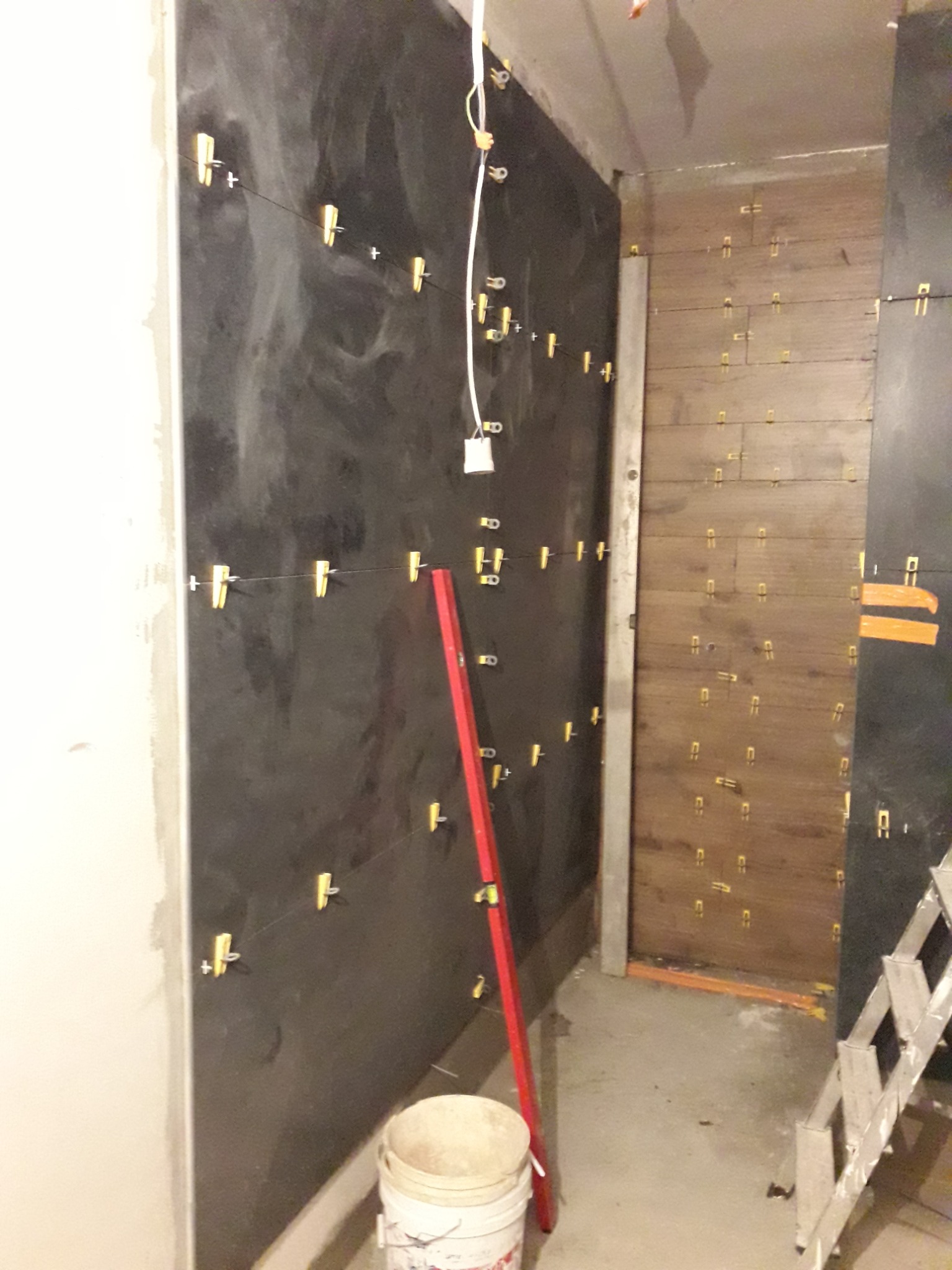 Ściany w trakcie układania płytek, widoczne poziomica, krzyżyki dystansowe i wiadro z zaprawą.