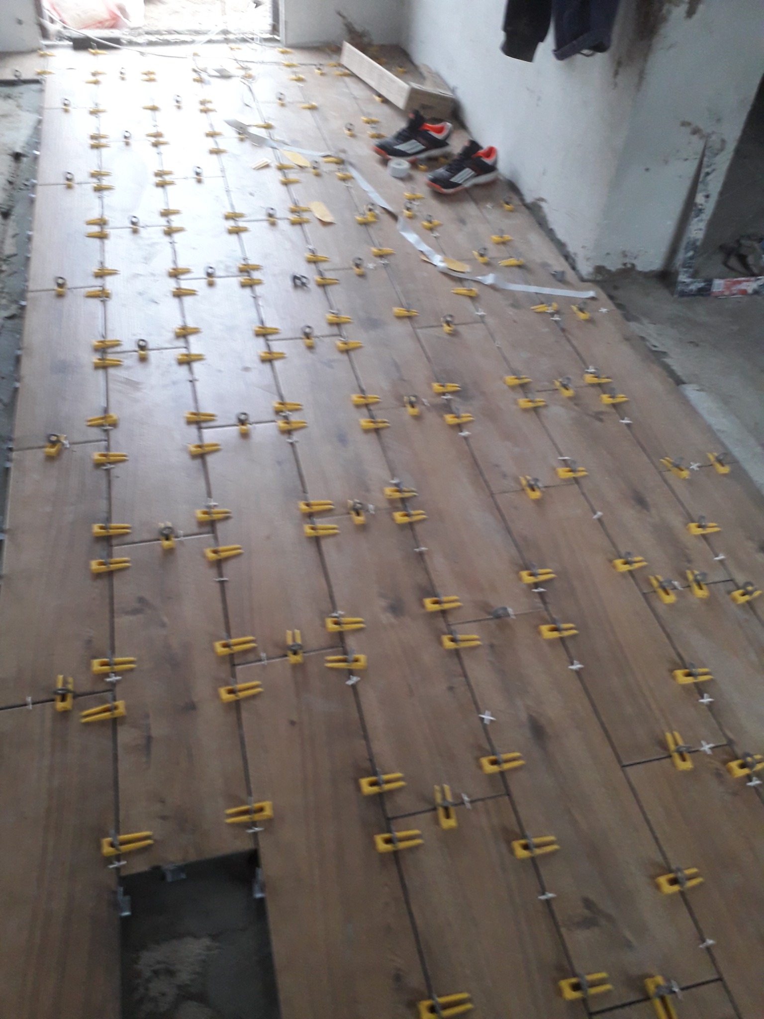 Układanie drewnopodobnych płytek podłogowych z użyciem klipsów poziomujących w trakcie prac w Suwałkach. W tle widoczne buty i narzędzia.