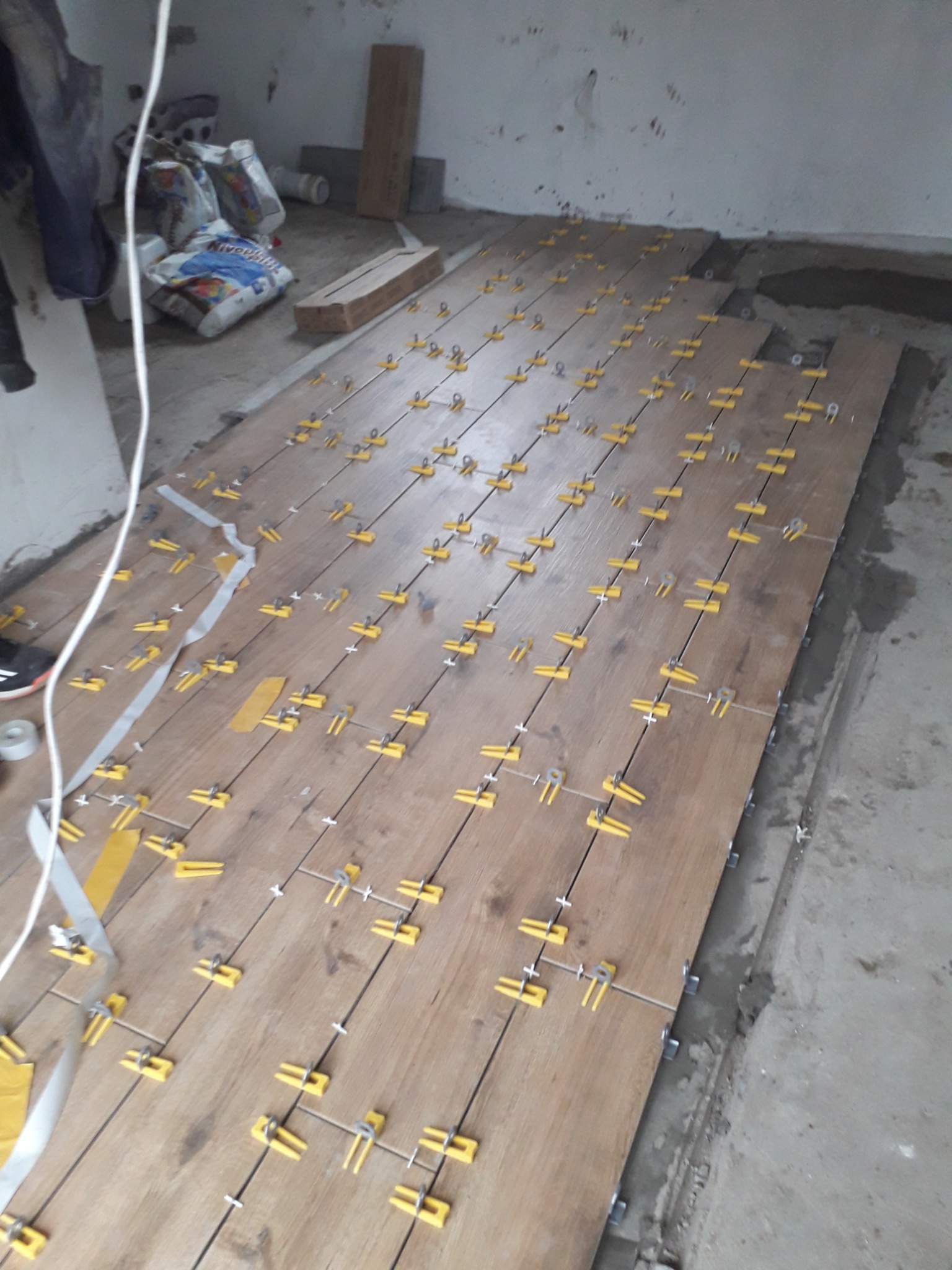 Układanie płytek imitujących drewno na podłodze, z zastosowaniem żółtych klipsów poziomujących, widoczne worki z zaprawą w tle.