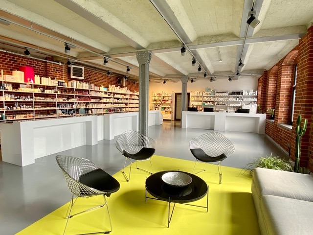 Przestronne wnętrze sklepu z kosmetykami: ceglane ściany, białe półki wypełnione produktami, nowoczesne krzesła druciane i żółty dywanik pod stolikiem kawowym.