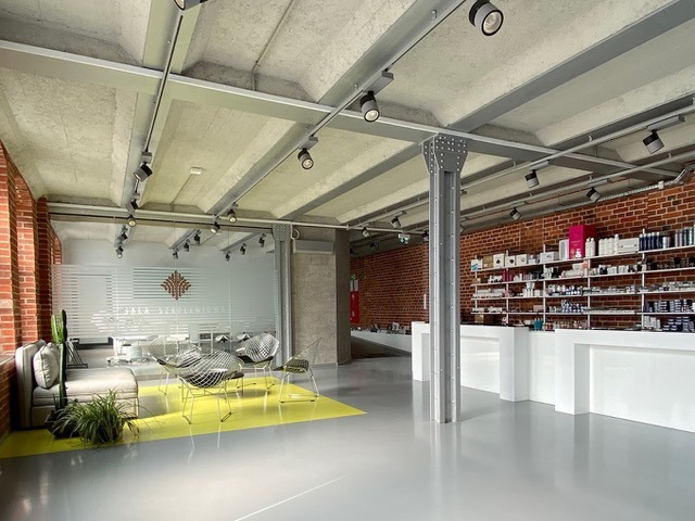 Wnętrze salonu kosmetycznego w stylu loft z ceglanymi ścianami, szarymi podłogami, betonowym sufitem z oświetleniem punktowym, przeszkloną salą zabiegową z napisem 'Sala Szkoleniowa' oraz regałami...