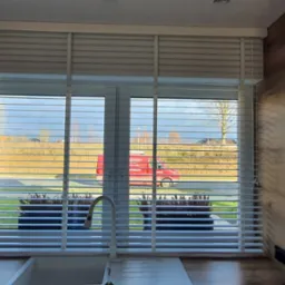 Białe żaluzje poziome w oknie kuchni, widok na zewnątrz z czerwonym samochodem serwisowym w tle.